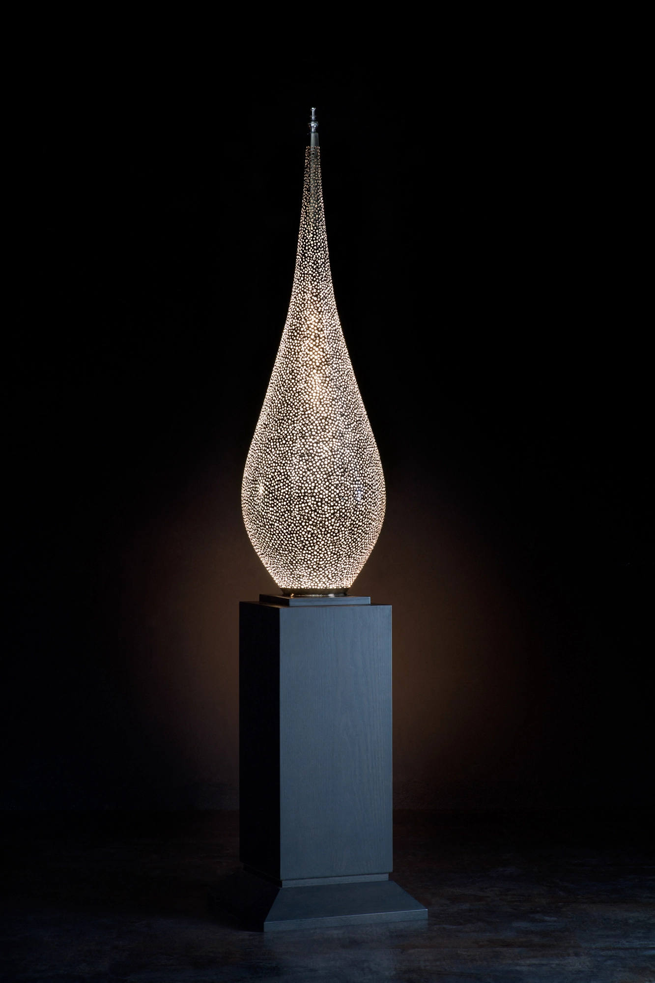 A slender pierced teardrop lamp glowing atop a tall pale pedestal, against a black background.