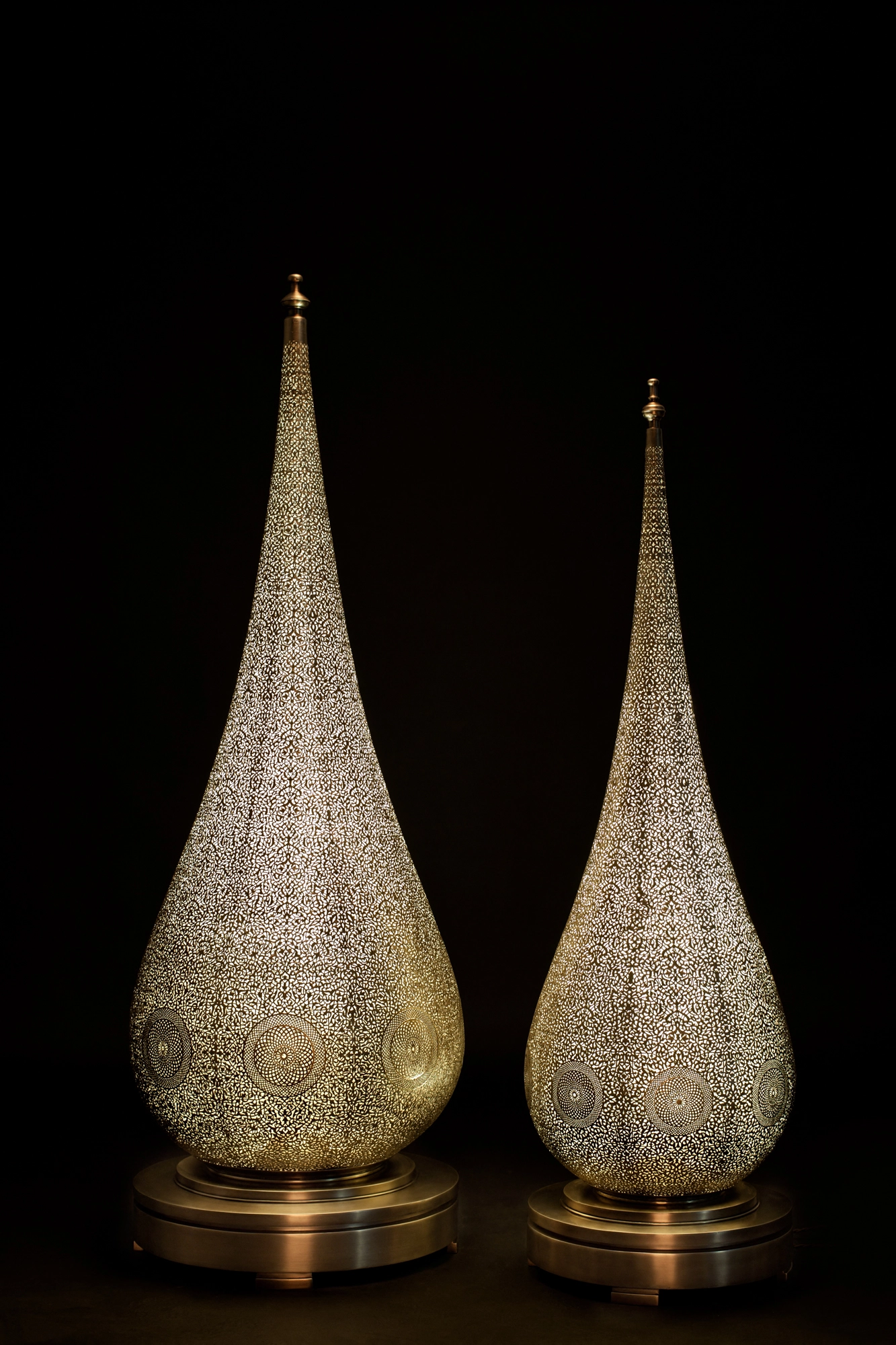 Two pierced brass teardrop floor lamps glowing side by side against a black background.