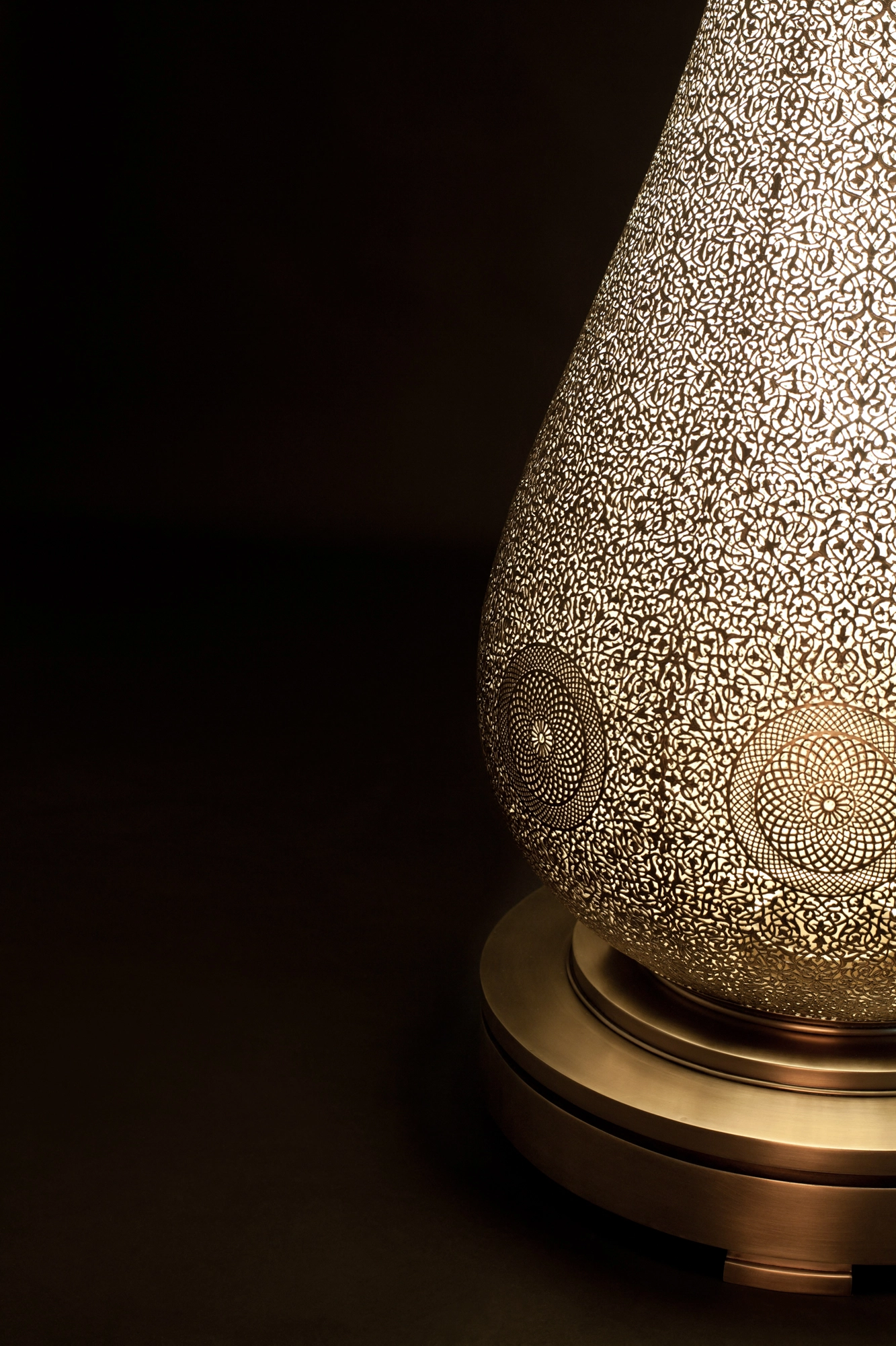 Close-up of a pierced teardrop floor lamp's lower body meeting its round brass base, glowing in the dark.