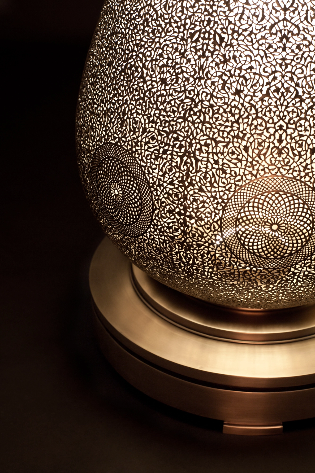 Close-up of a pierced floor lamp's glowing body and tiered round brass base against a dark background.