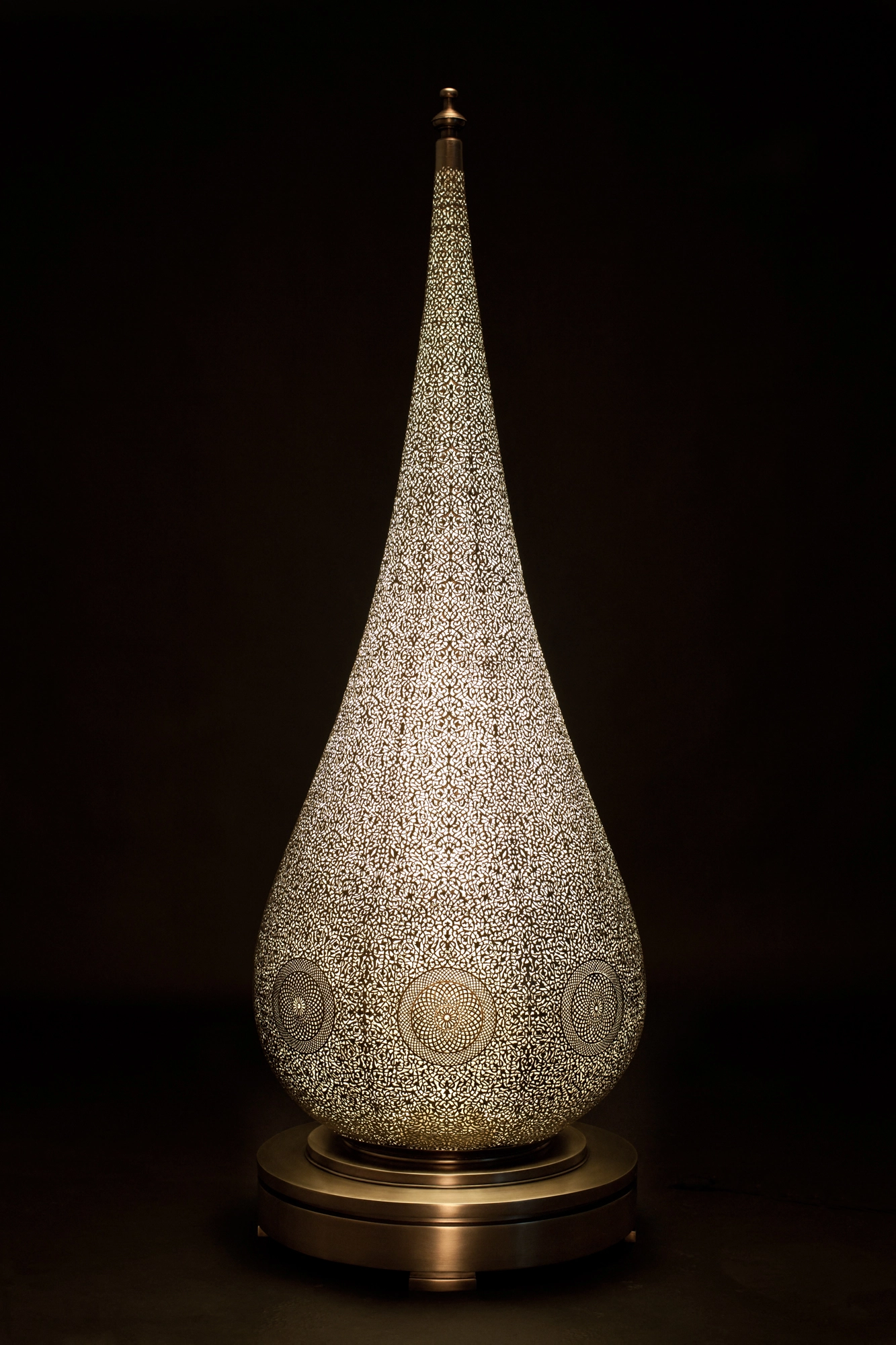 A pierced teardrop floor lamp with a pointed finial on a round brass base, against a black background.
