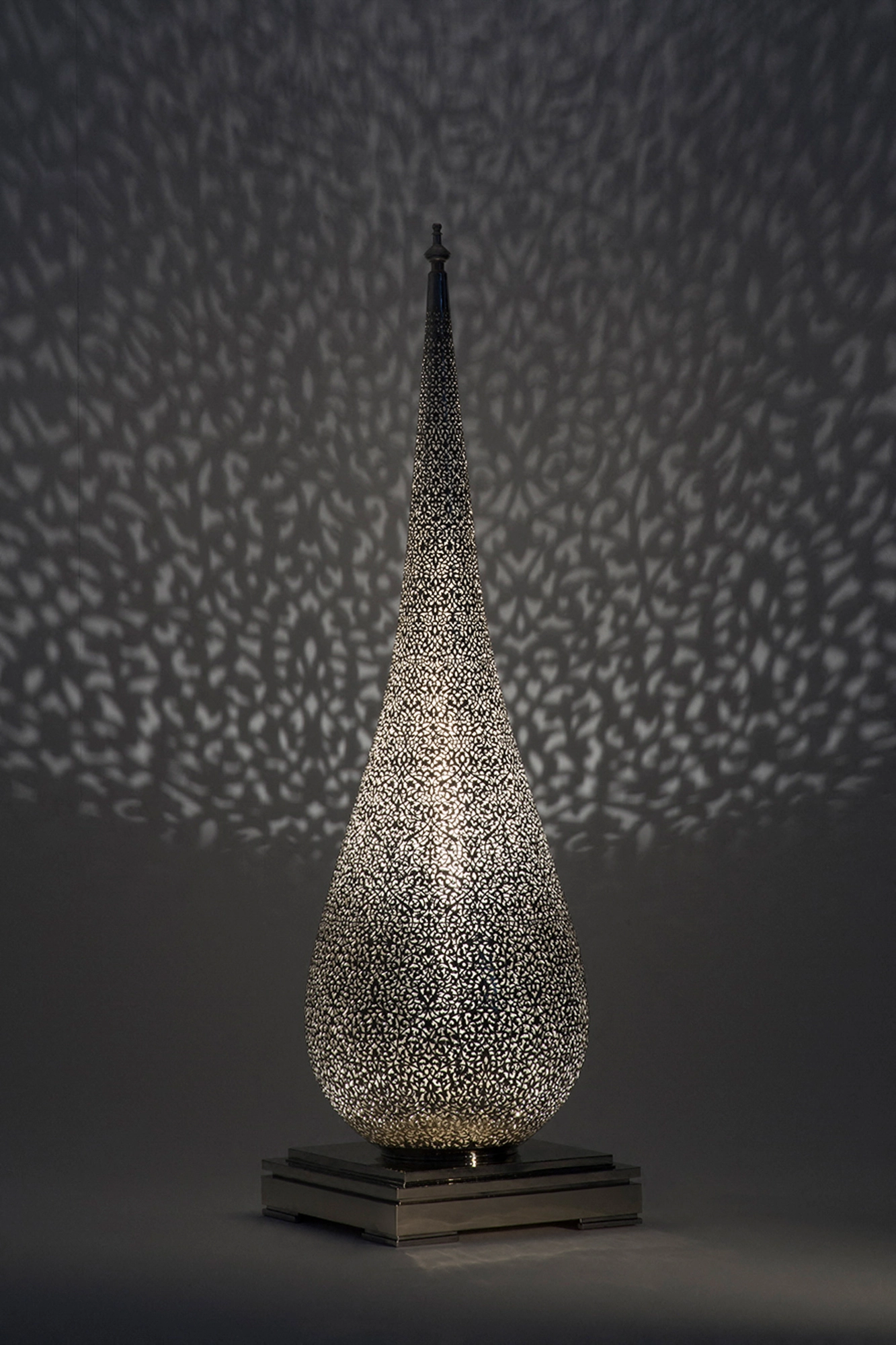 A tall narrow pierced teardrop floor lamp on a square base, scattering a fine dotted spray of light.