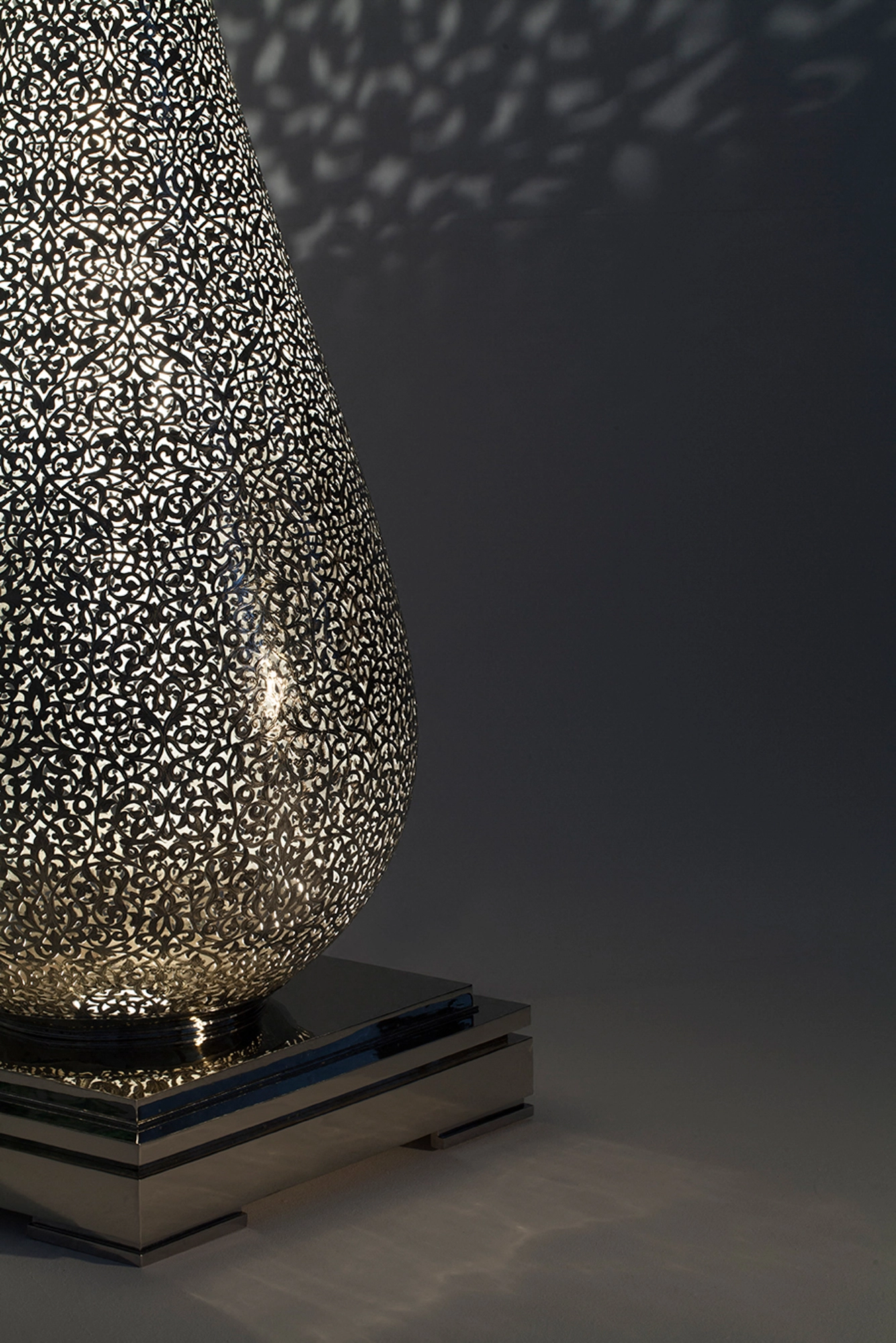 An angled view of a pierced teardrop floor lamp's body glowing on its base, dotted light around it.