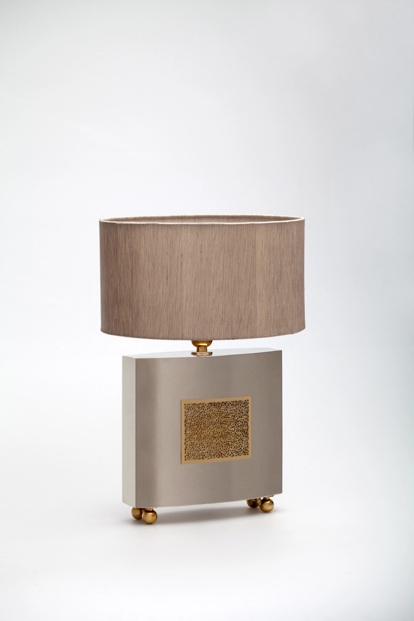 Table lamp with a beige drum shade on a brushed-metal base inset with a small gilded floral panel, on white.