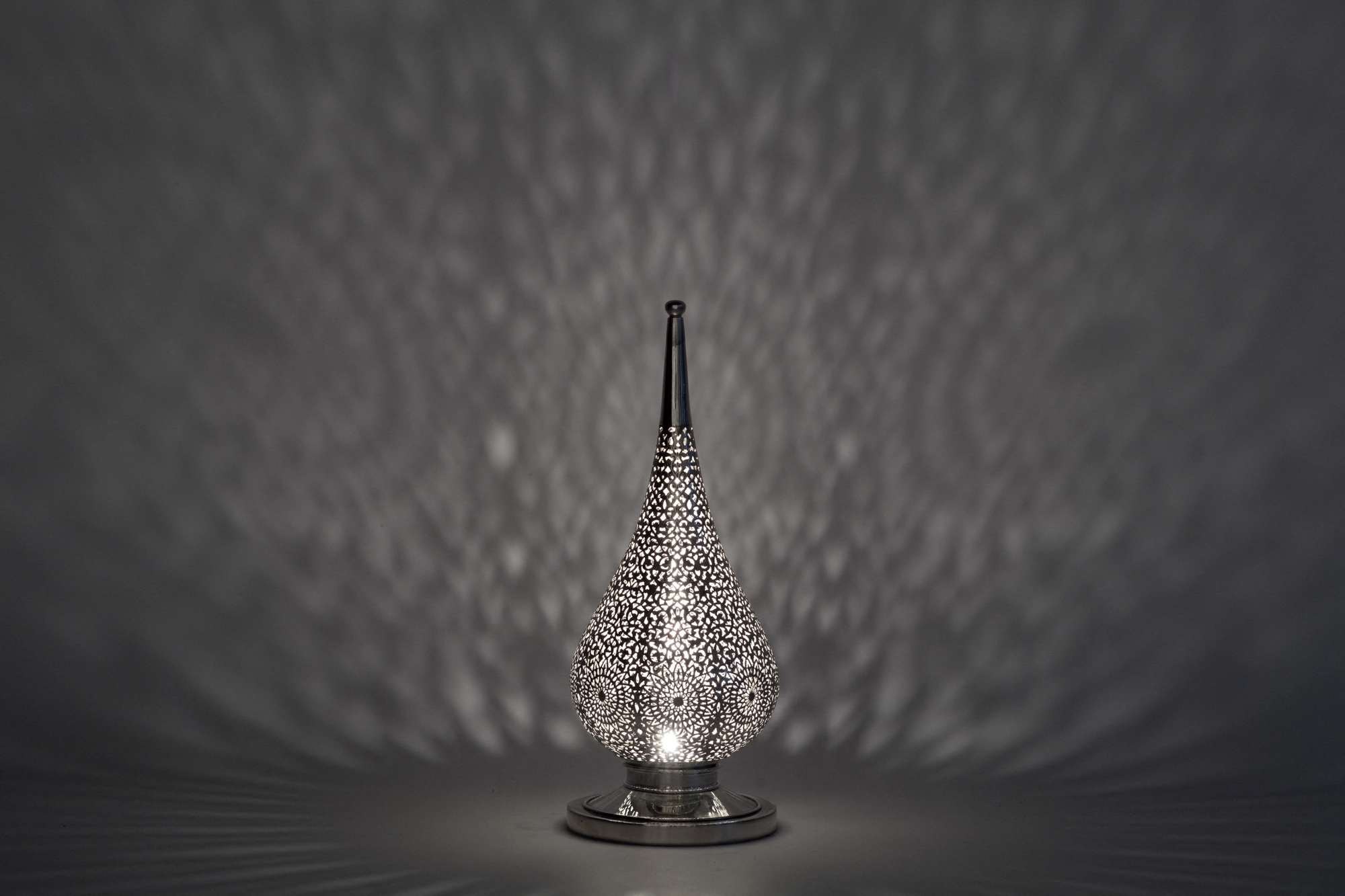 A small flame-shaped pierced candle holder casting faint dappled light against a soft grey background.