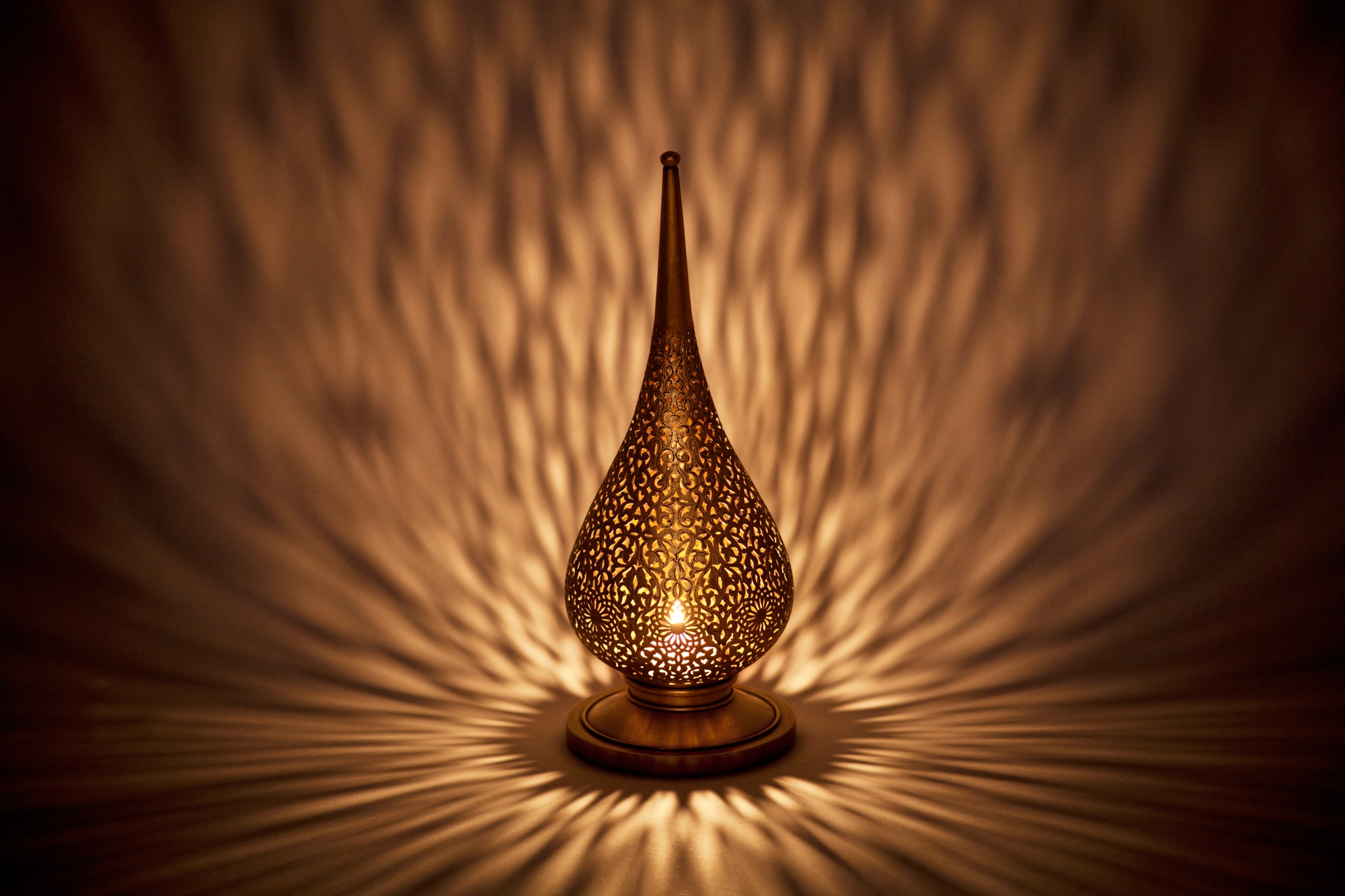 A small flame-shaped pierced candle holder casting a warm orange burst of dappled light around its base.