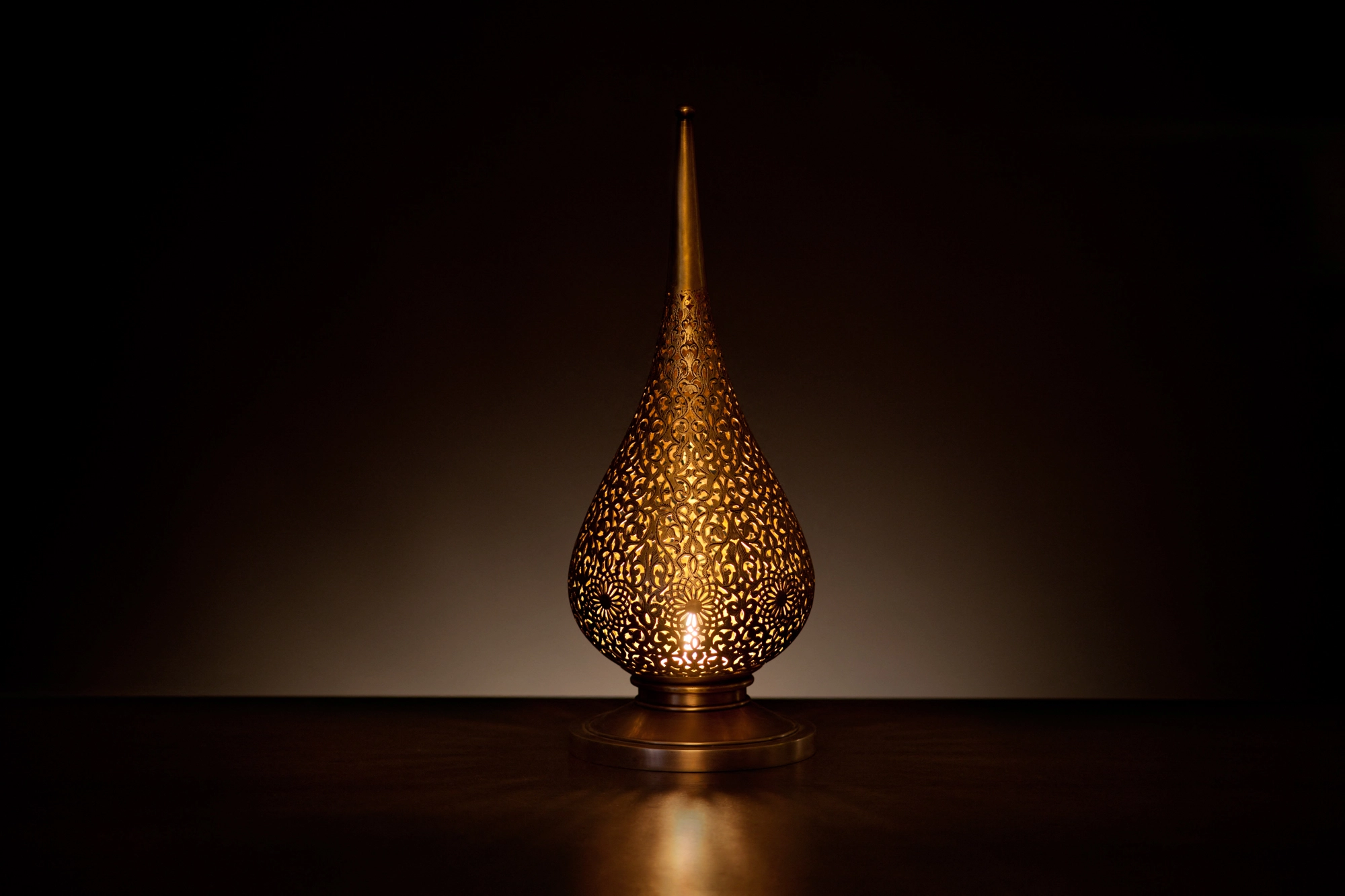 A small flame-shaped pierced candle holder glowing warmly with its reflection on a dark surface.