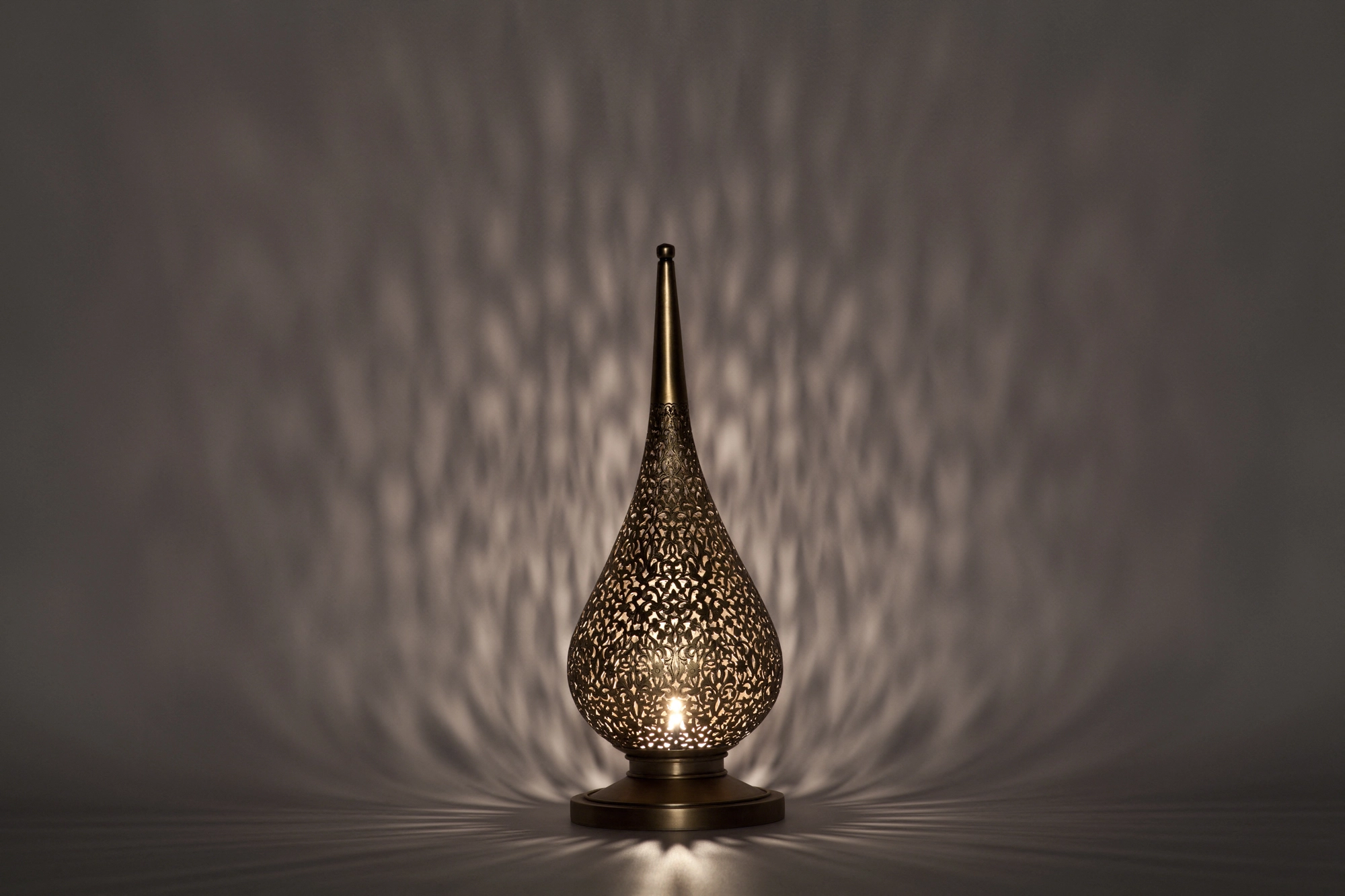 A small flame-shaped pierced candle holder scattering soft dappled light across a grey background.