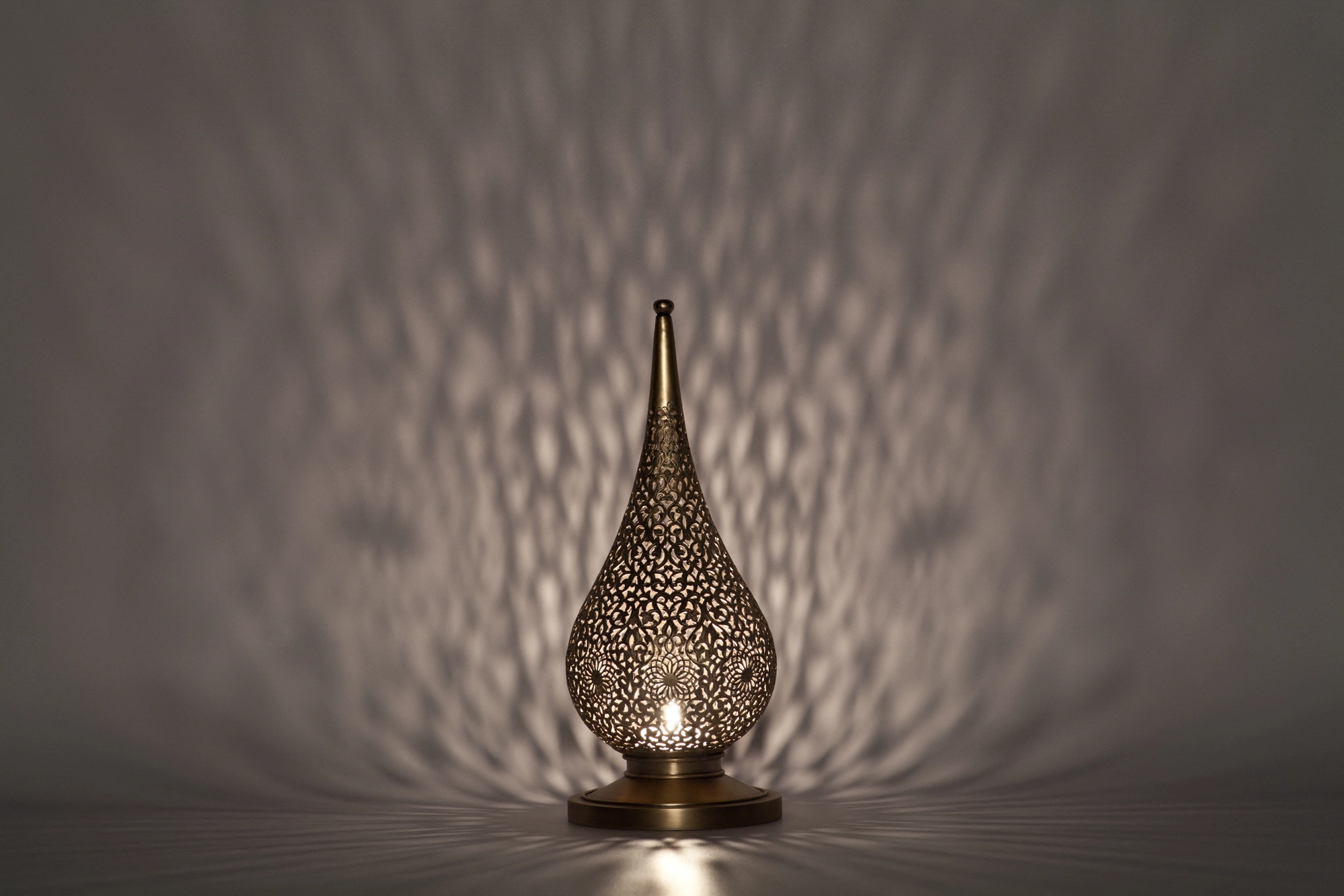 A small flame-shaped pierced metal candle holder casting a fan of dappled shadow on a grey background.