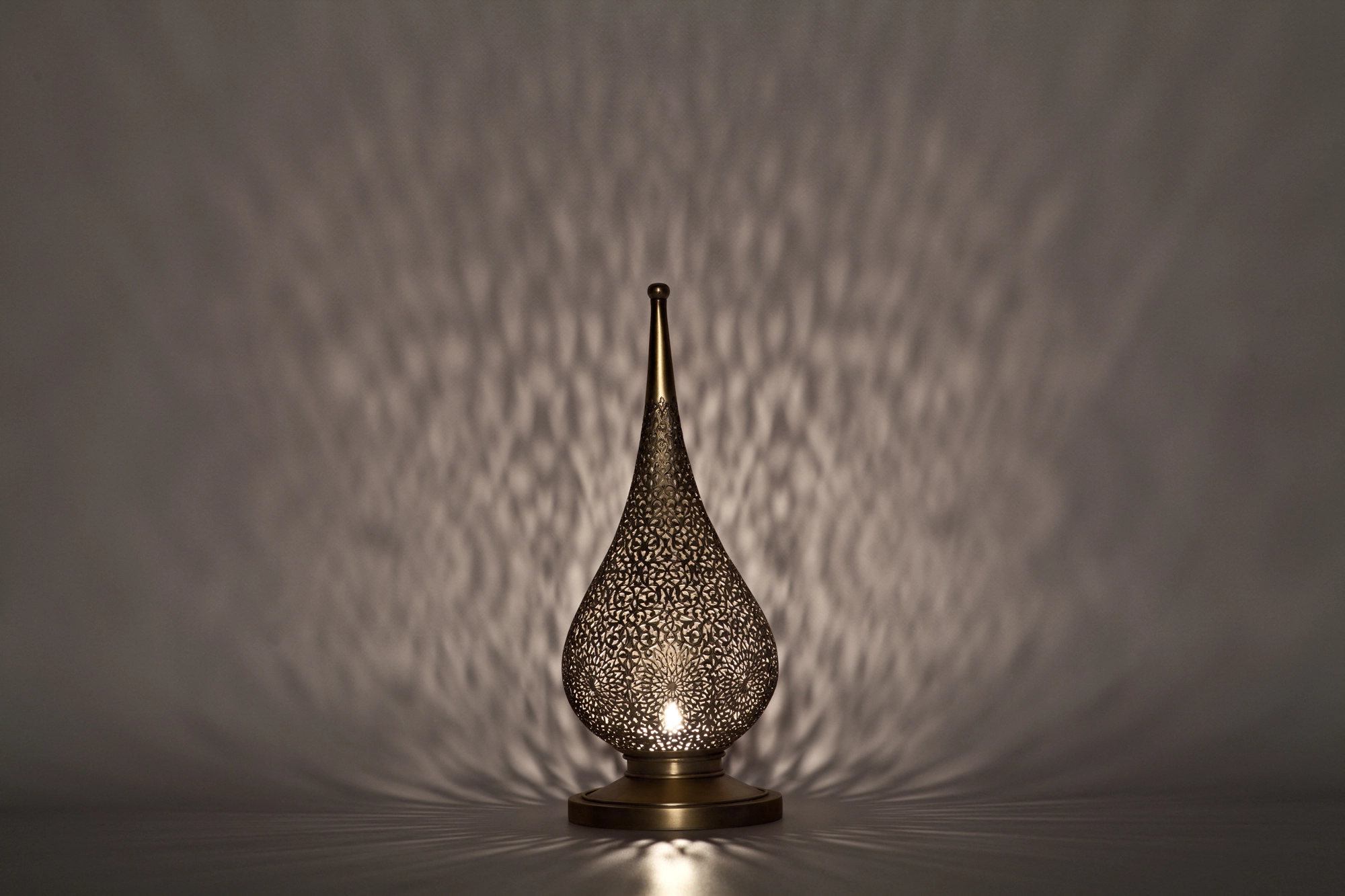 A small flame-shaped pierced candle holder lit from within, casting pale dappled light on a light grey background.