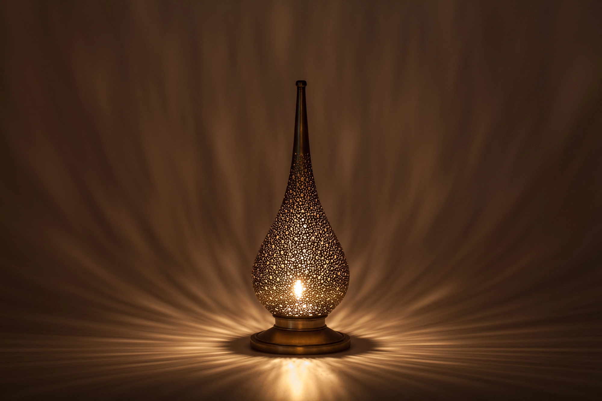 A small flame-shaped pierced candle holder glowing warmly above its reflection on a dark surface.