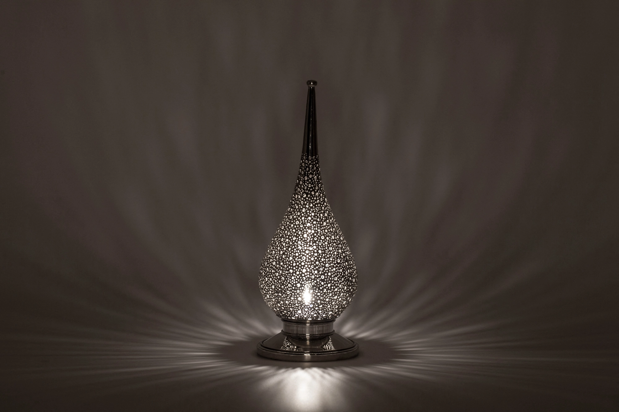 A small flame-shaped pierced candle holder casting soft dappled light above its reflection on a grey surface.