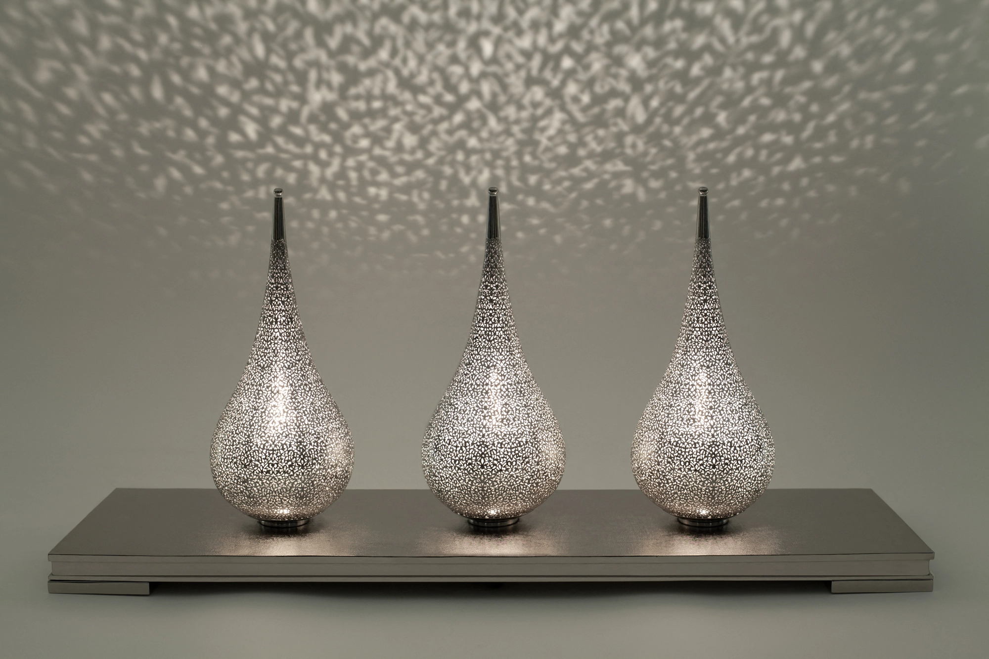 Three pierced teardrop lamps standing on a single tray base, scattering dotted light against a grey wall.