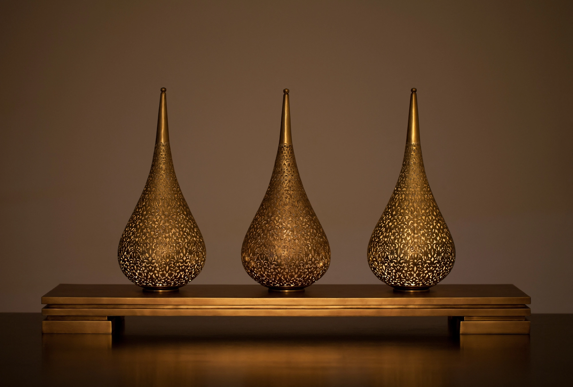 Three pierced teardrop lamps glowing warmly in a row on a low wooden shelf, lit from within.