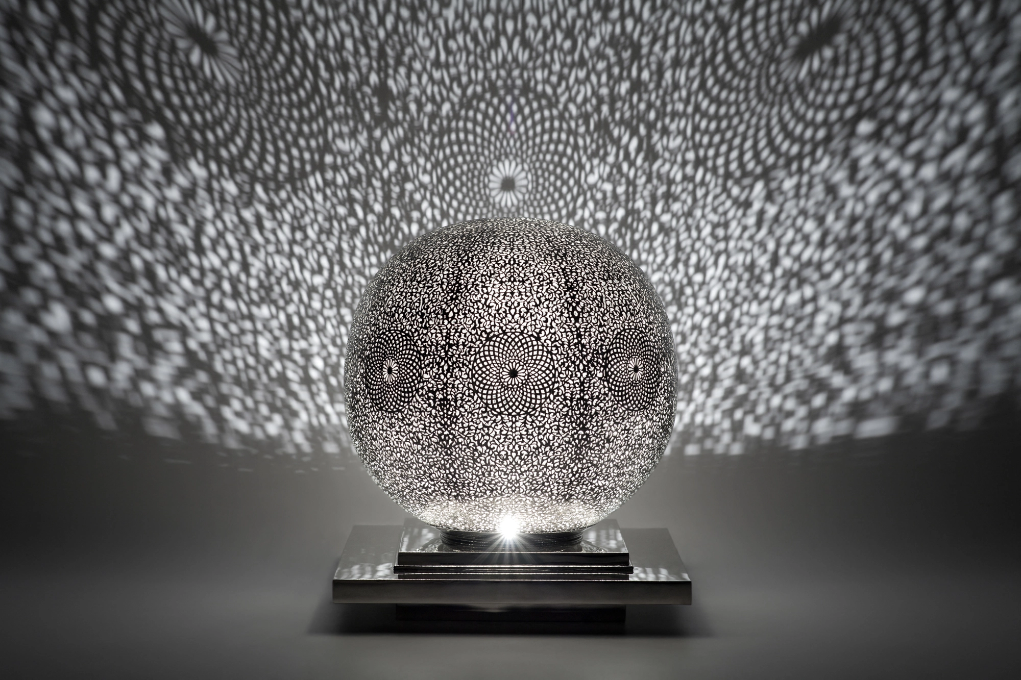 A spherical pierced metal candle holder lit from within, scattering fine dappled light across a grey background.