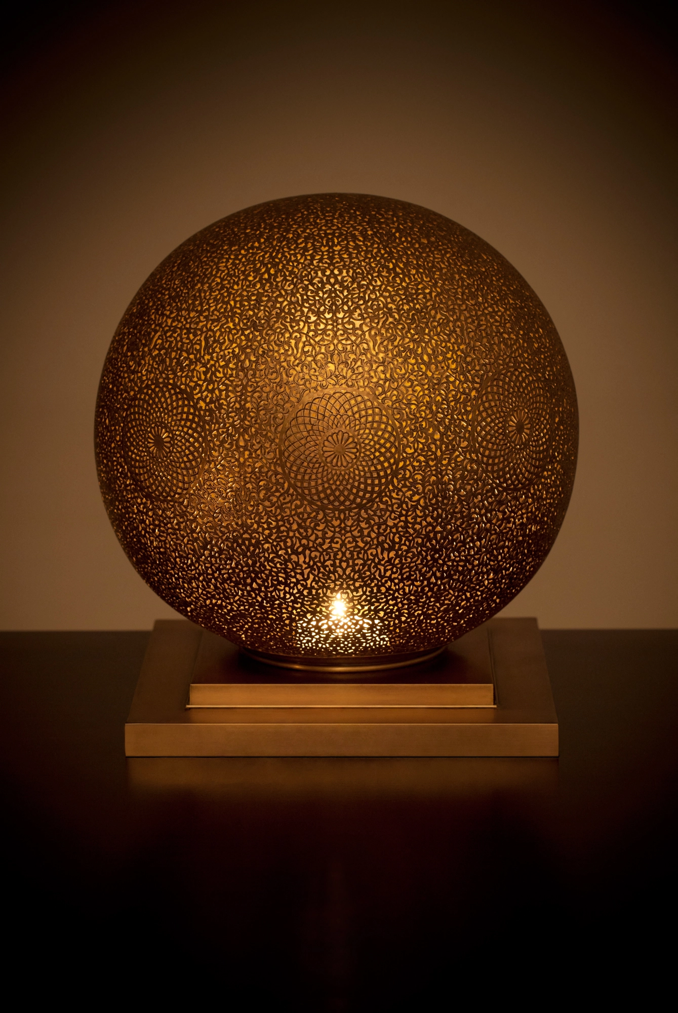 A spherical pierced metal candle holder glowing amber on a wooden base against a dark background.