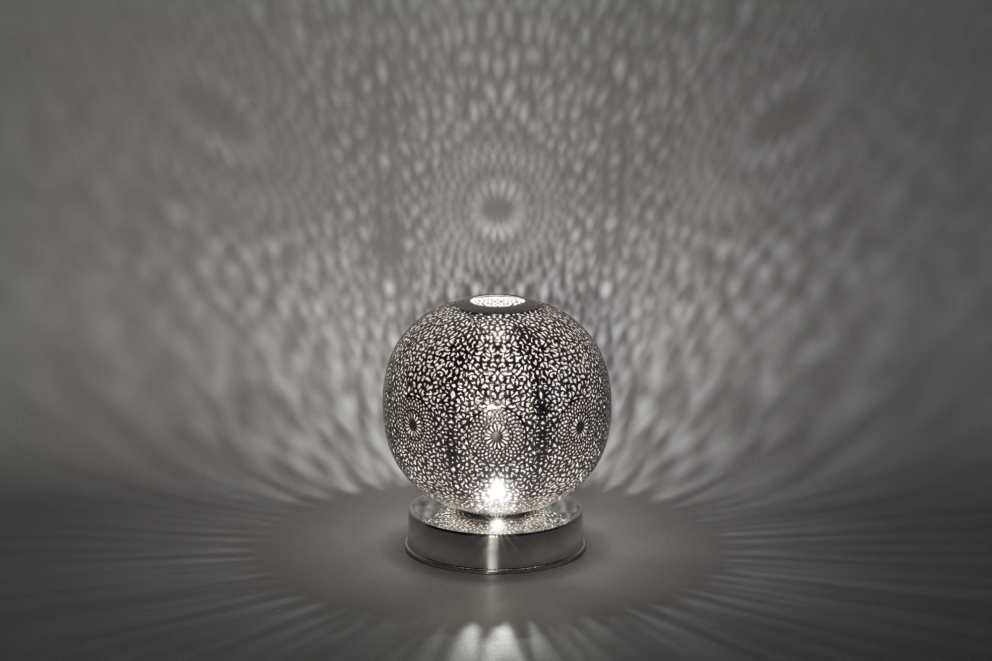 A small spherical pierced candle holder scattering fine dappled light above its reflection on a grey surface.