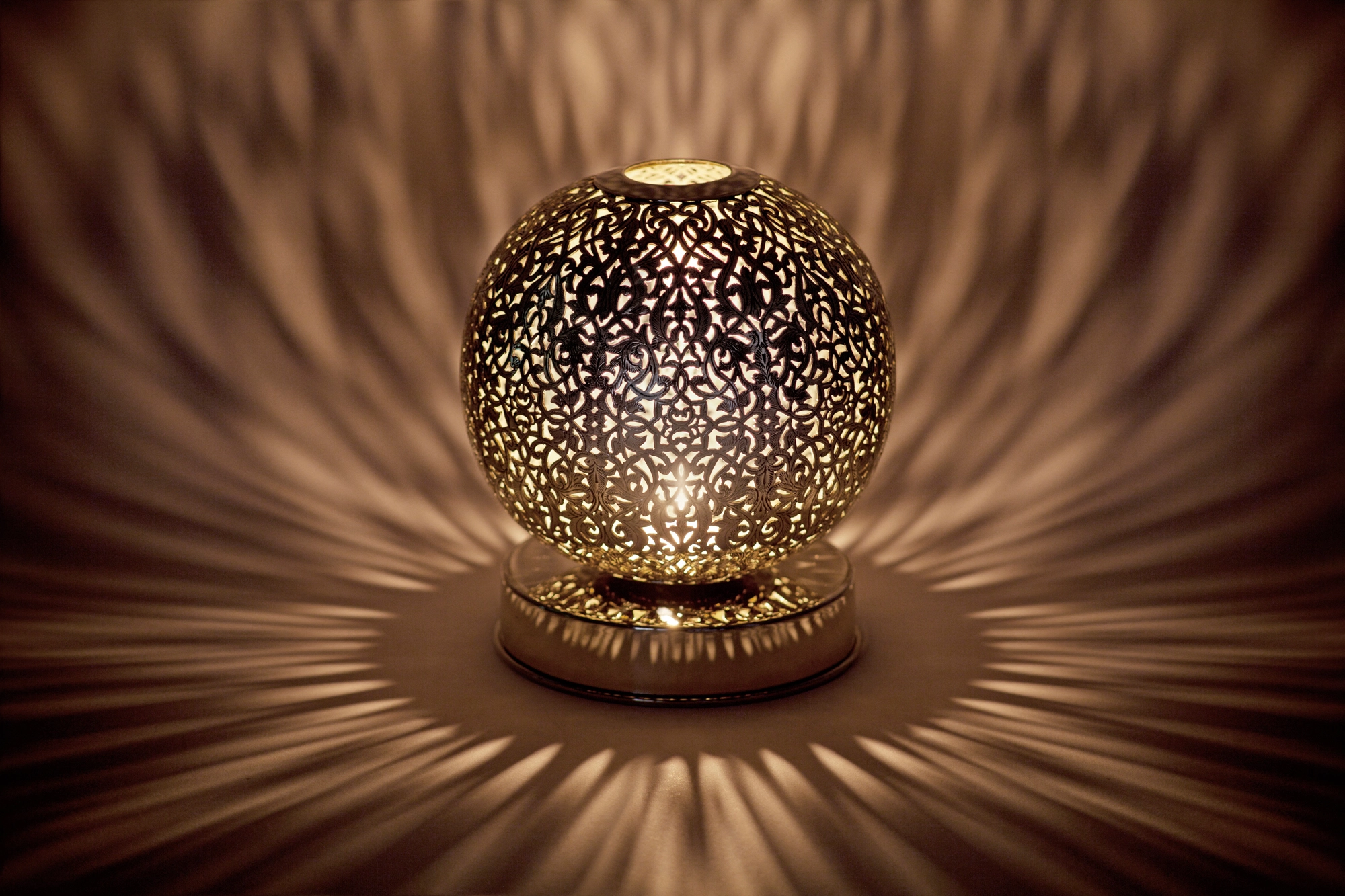 A small spherical pierced candle holder casting a warm burst of dappled light across a dark surface.