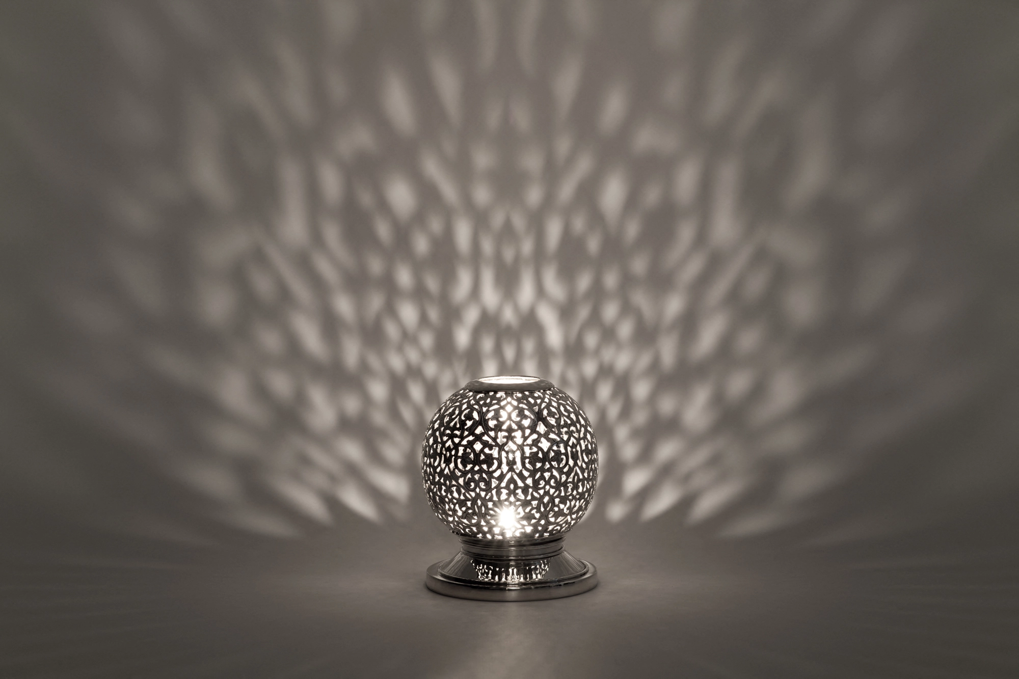 A small spherical pierced metal candle holder casting soft dappled light on a grey background.