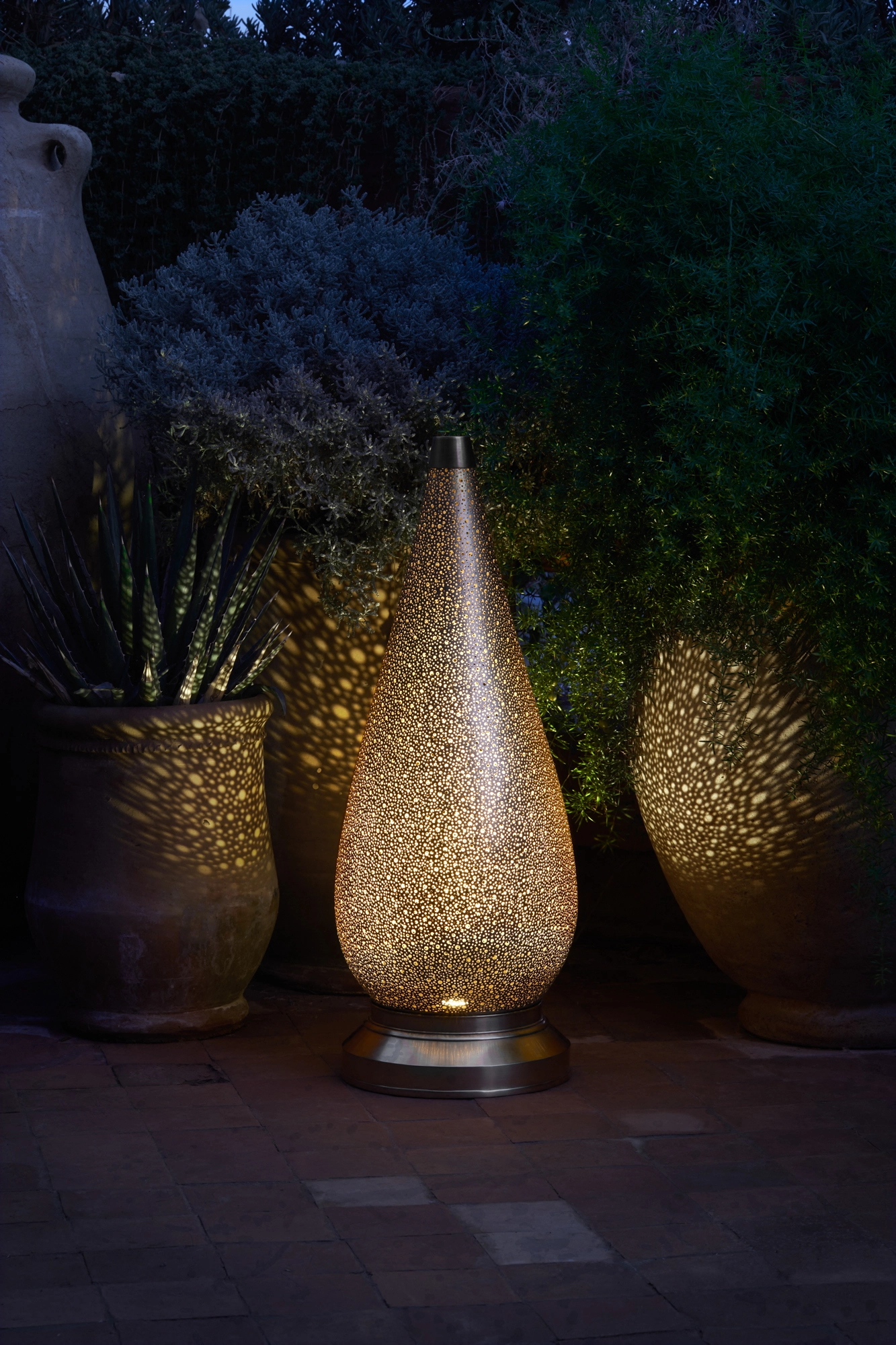 A pierced teardrop floor lamp glowing among garden planters at night, lighting the surrounding foliage.