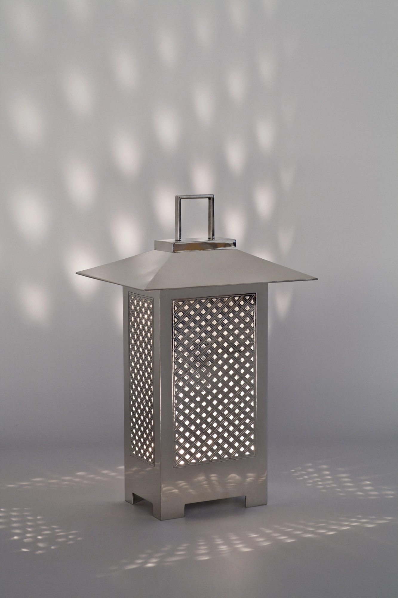 A silver lantern with a pagoda-style roof and criss-cross lattice panels, scattering dotted light on a grey wall.