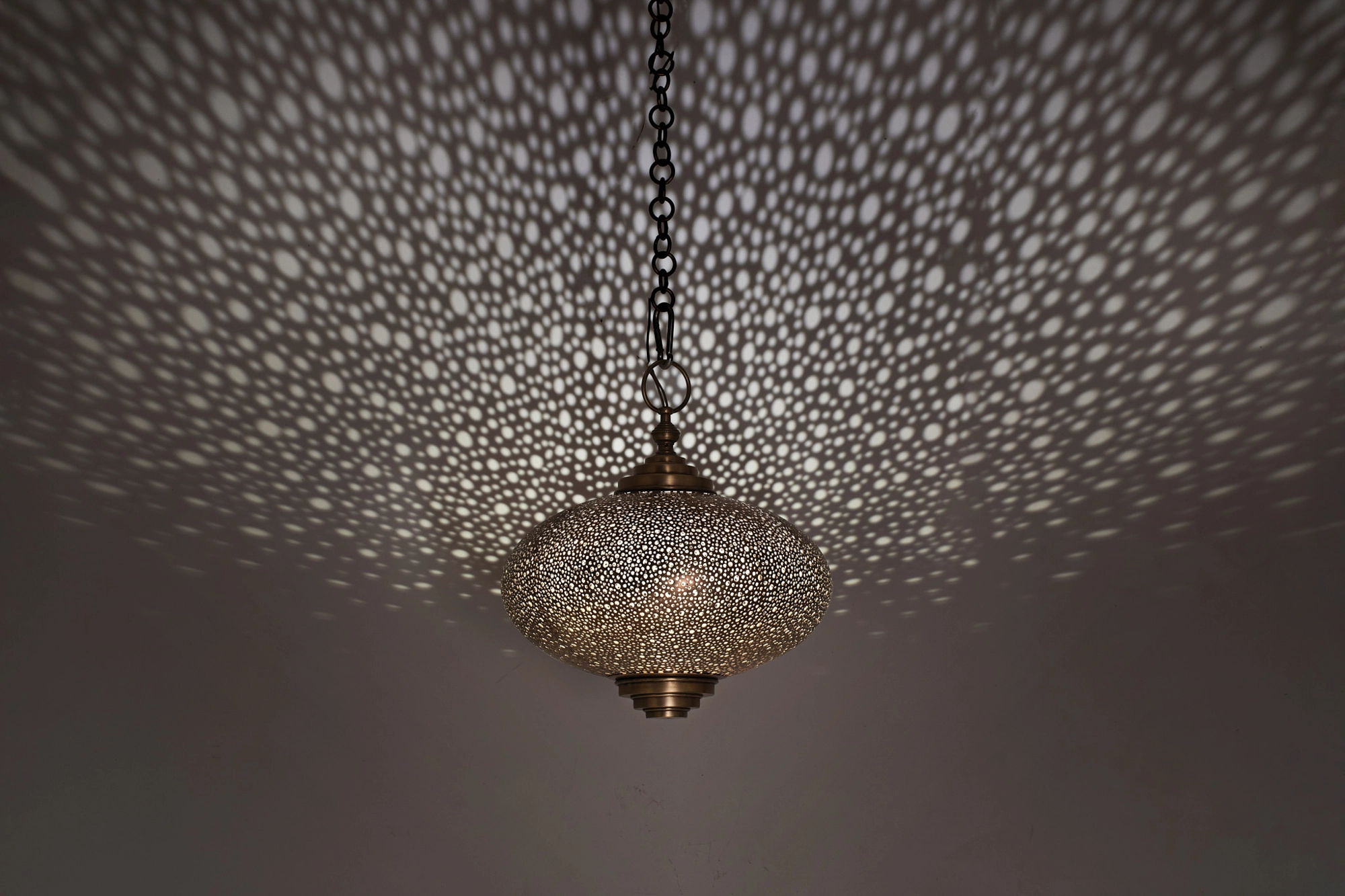 Pierced squashed-globe pendant seen from below, glowing amid a wide halo of light dots on the ceiling.