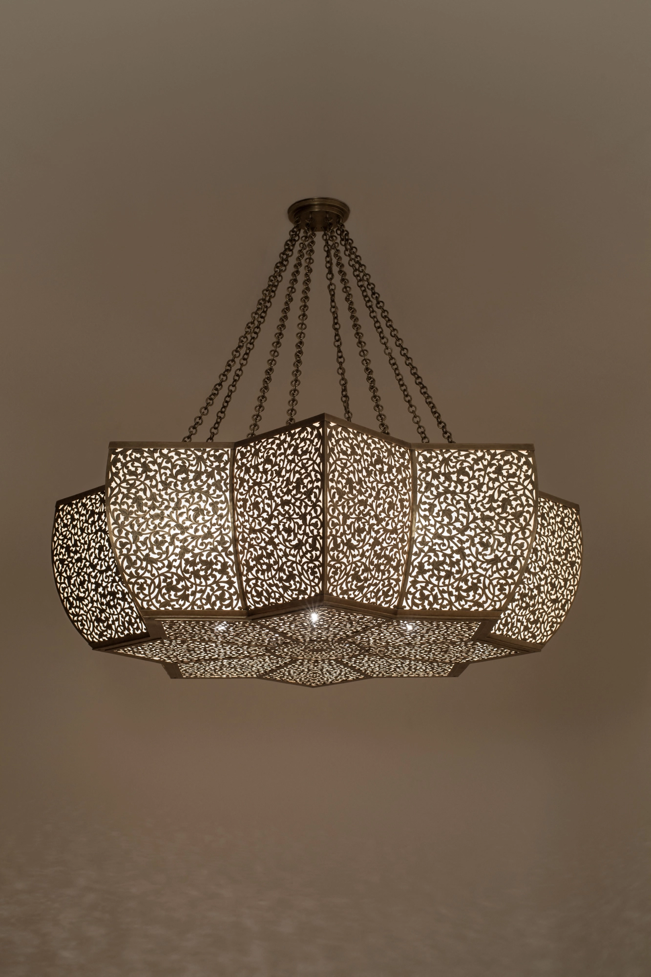 A wide pierced octagonal chandelier with flower-like lobes, suspended on chains in a dim room.