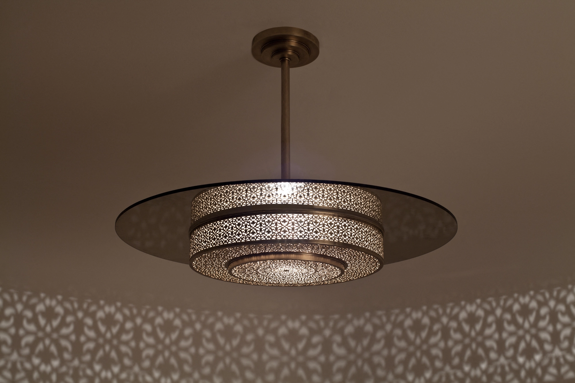 Stacked pierced-brass disc-drum pendant on a short stem, glowing softly beneath a patterned ceiling.