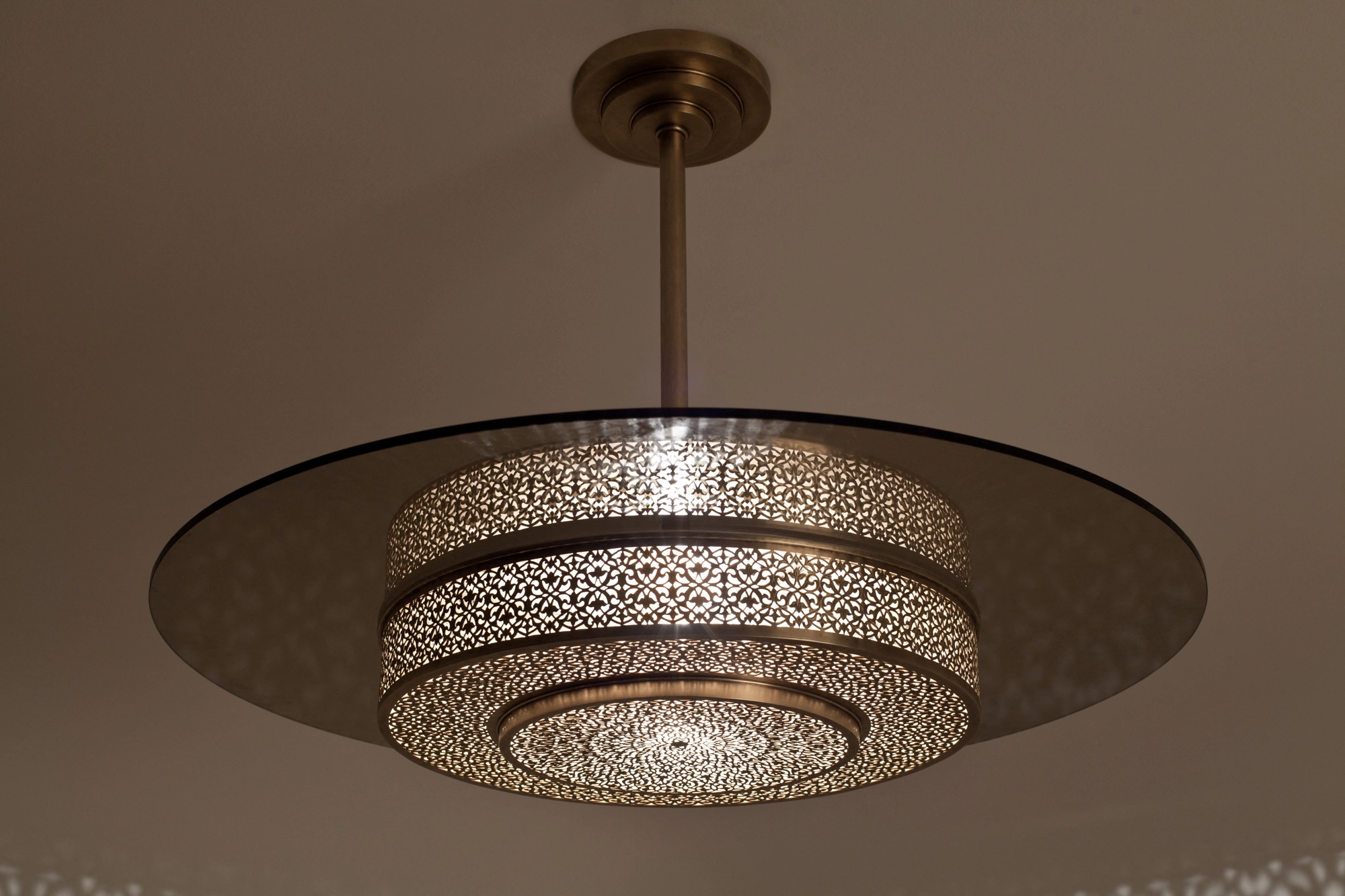 Tiered pierced-brass disc-drum pendant suspended from a stem, lit warmly against a soft-patterned ceiling.