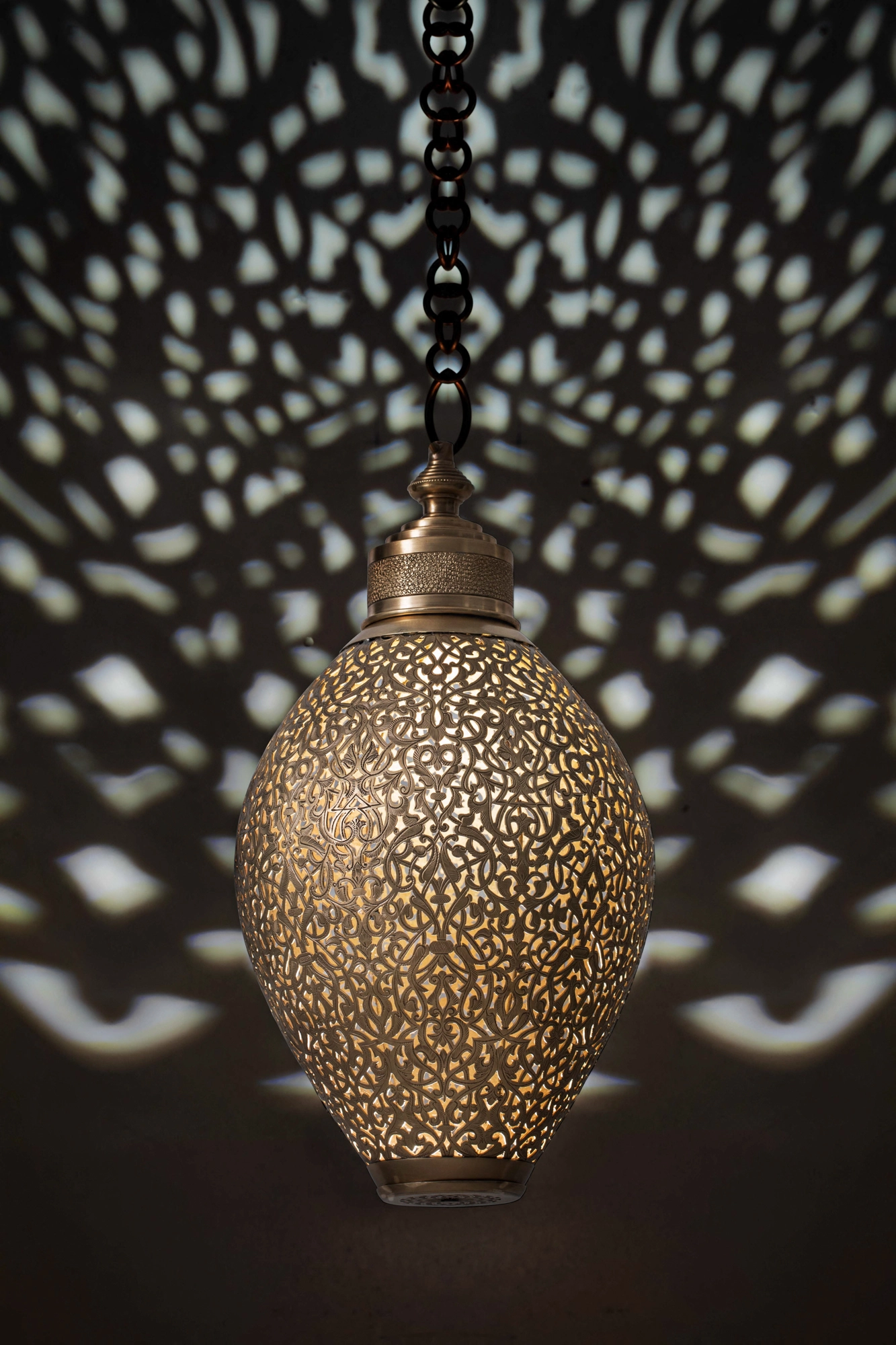 Ovoid pierced-brass amphora pendant lamp on a chain, casting a wide halo of pierced light dots across a dark wall.