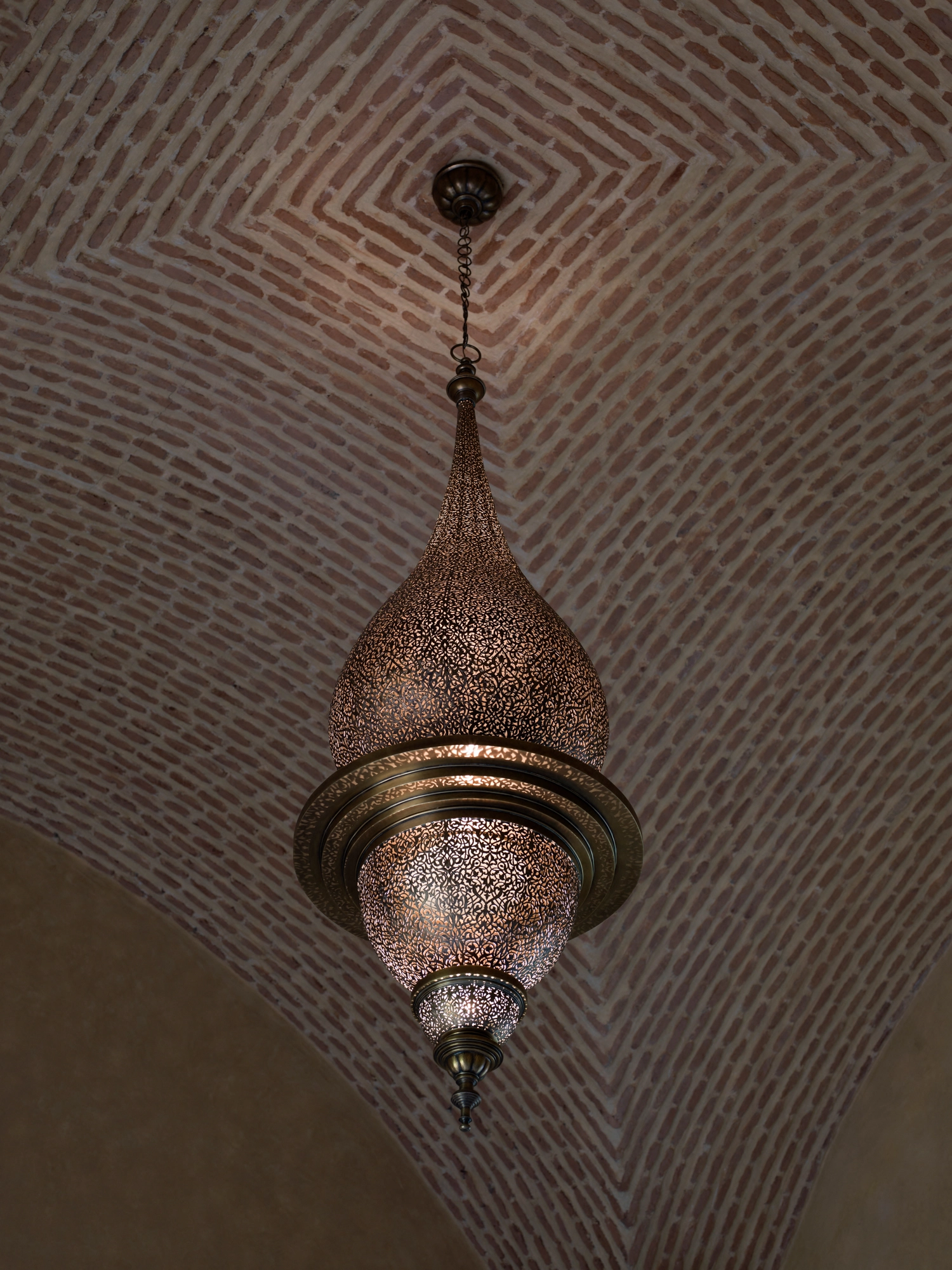 A small pierced teardrop pendant lamp hanging from a vaulted brick ceiling, glowing softly.