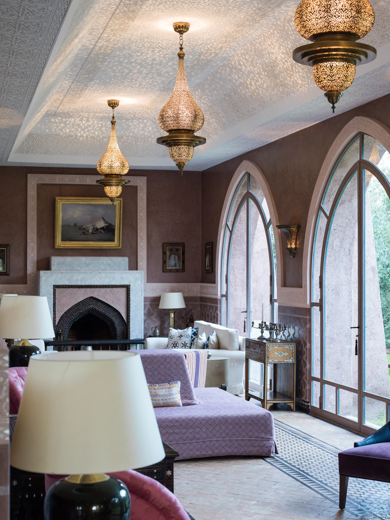 Mauve sitting room with a fireplace, table lamp and two pierced hanging lanterns by arched windows.