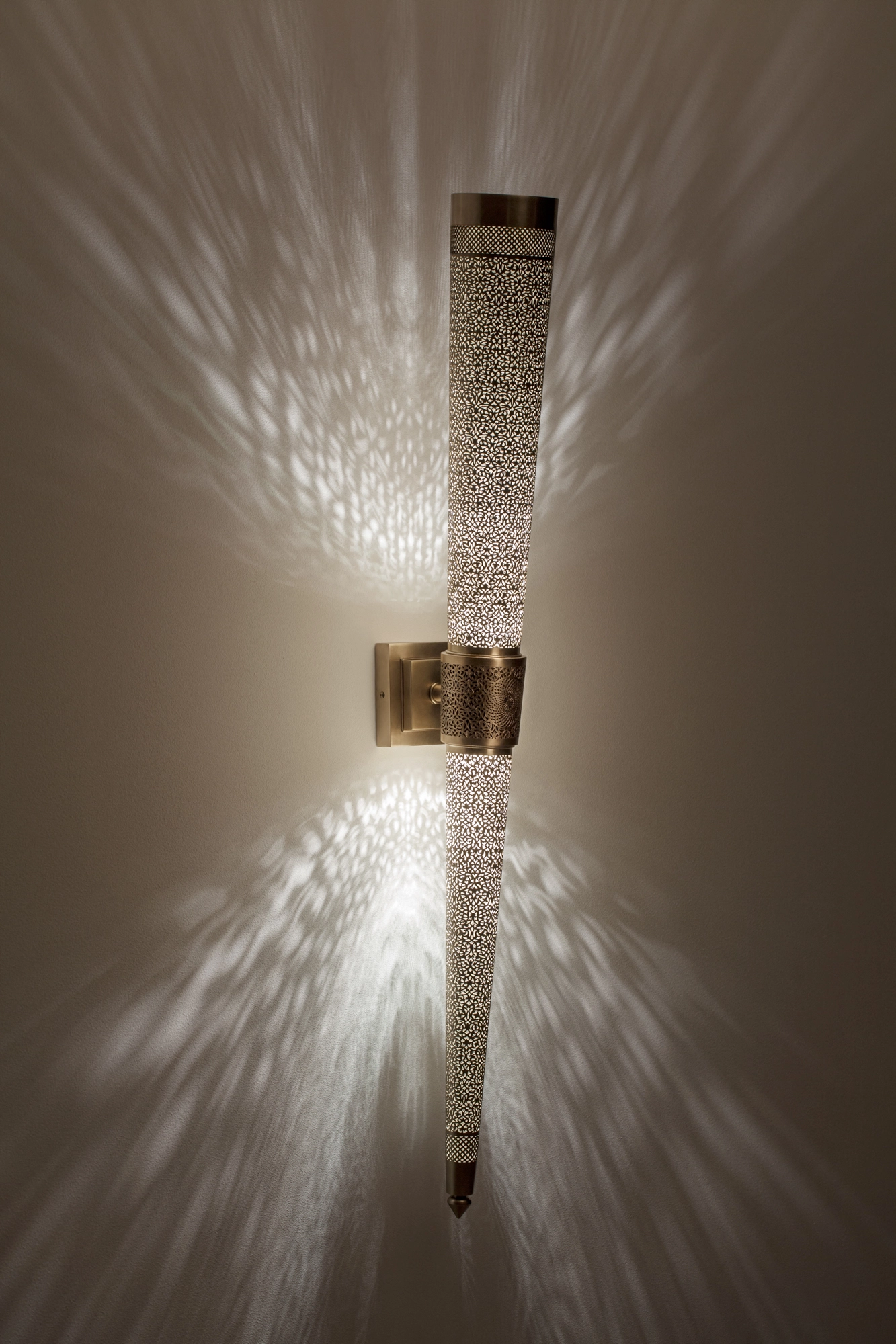 Slim tapering pierced-brass torch wall light on a bracket, casting a tall symmetrical burst of light up and down.