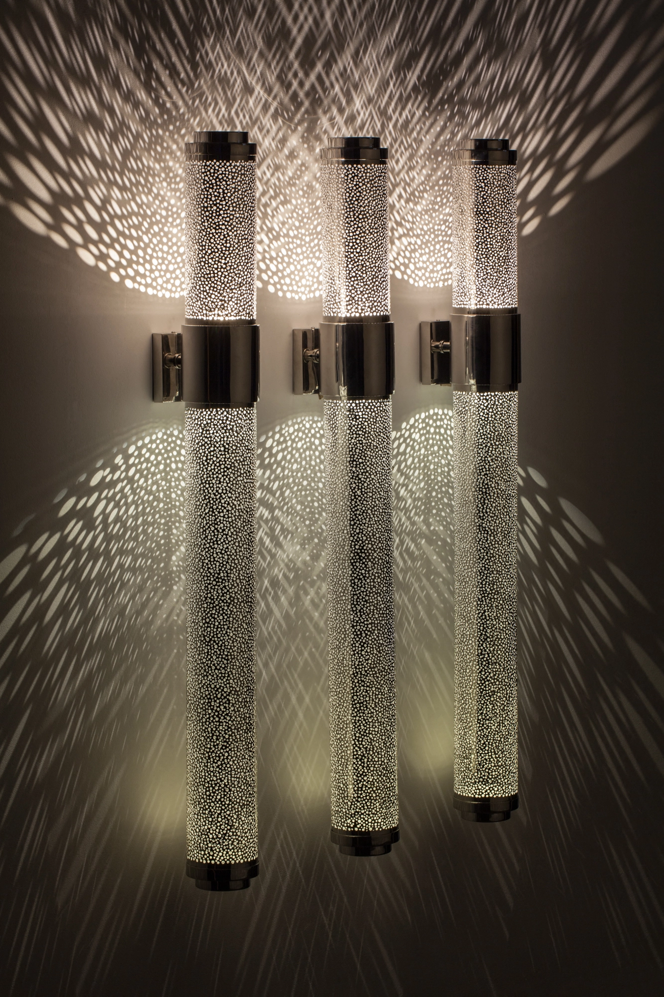 Cluster of pierced-brass cylinders at offset heights forming a wall light, scattering a wide feathered light burst.