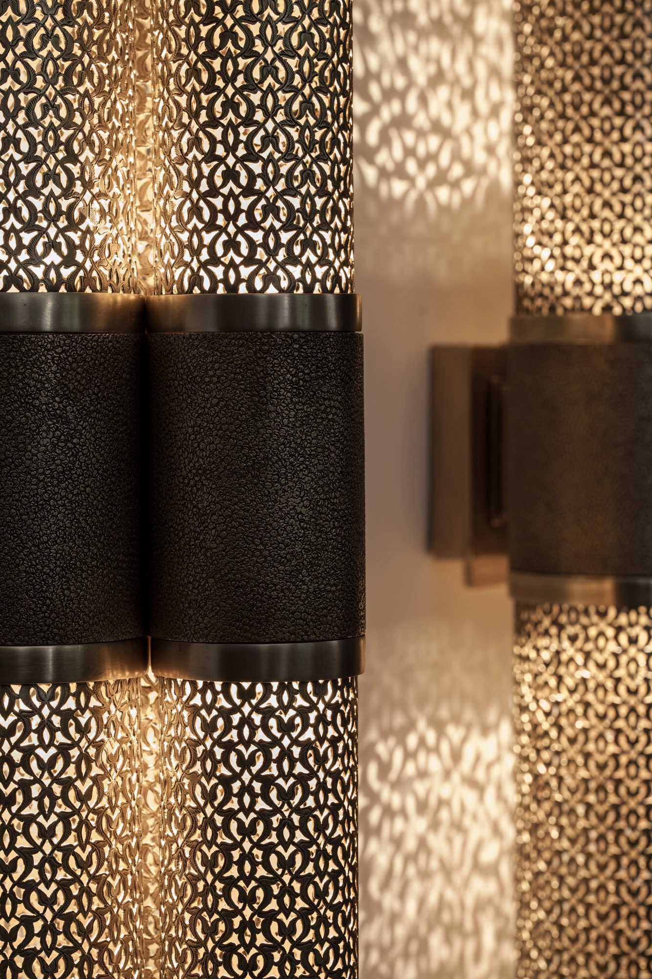 Close-up of pierced-brass triple-cylinder wall lights, dark bands dividing their glowing fretwork surfaces.