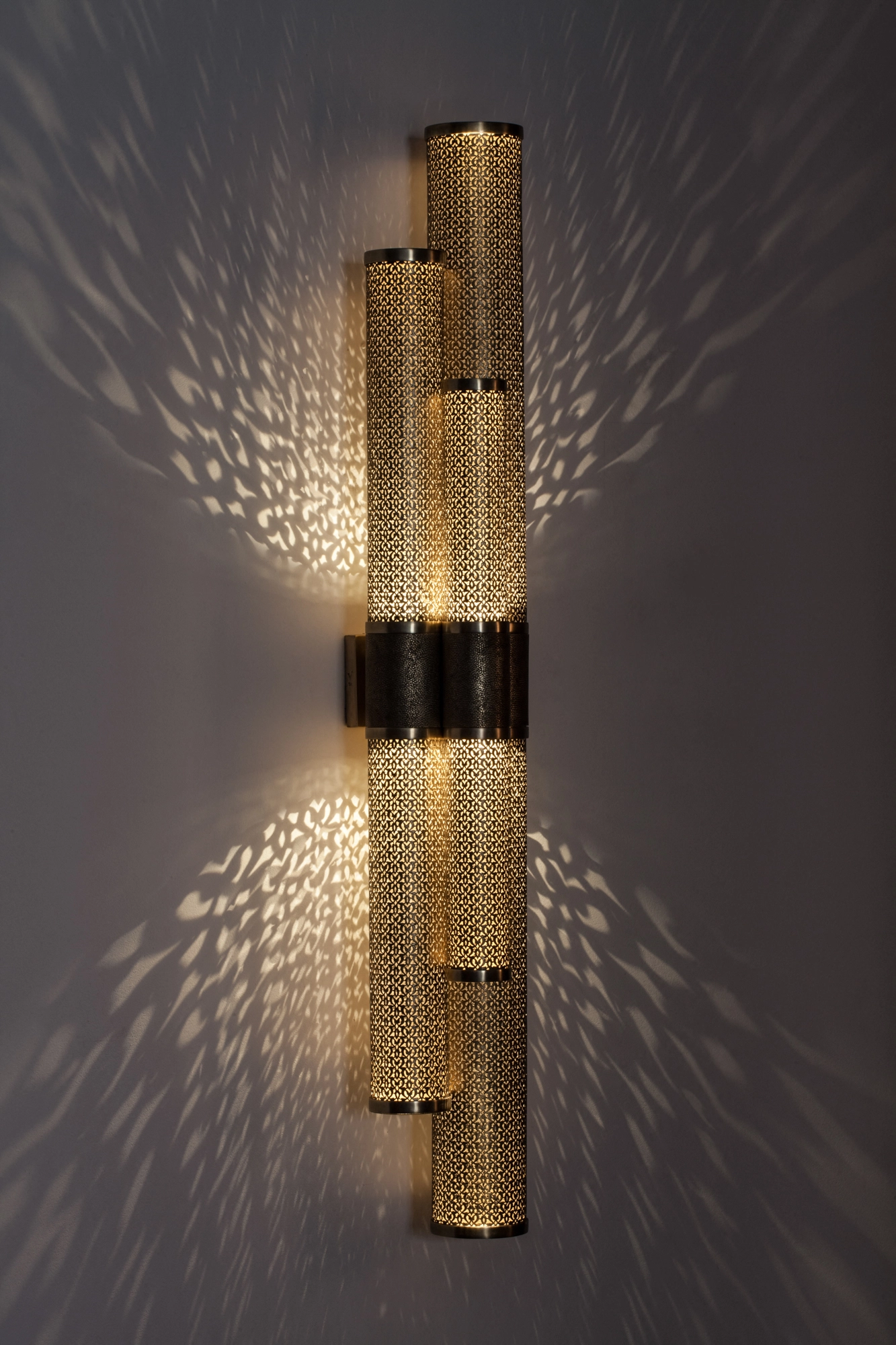 Cluster of three pierced-brass cylinders forming a vertical wall light, glowing with a soft feathered light spray.