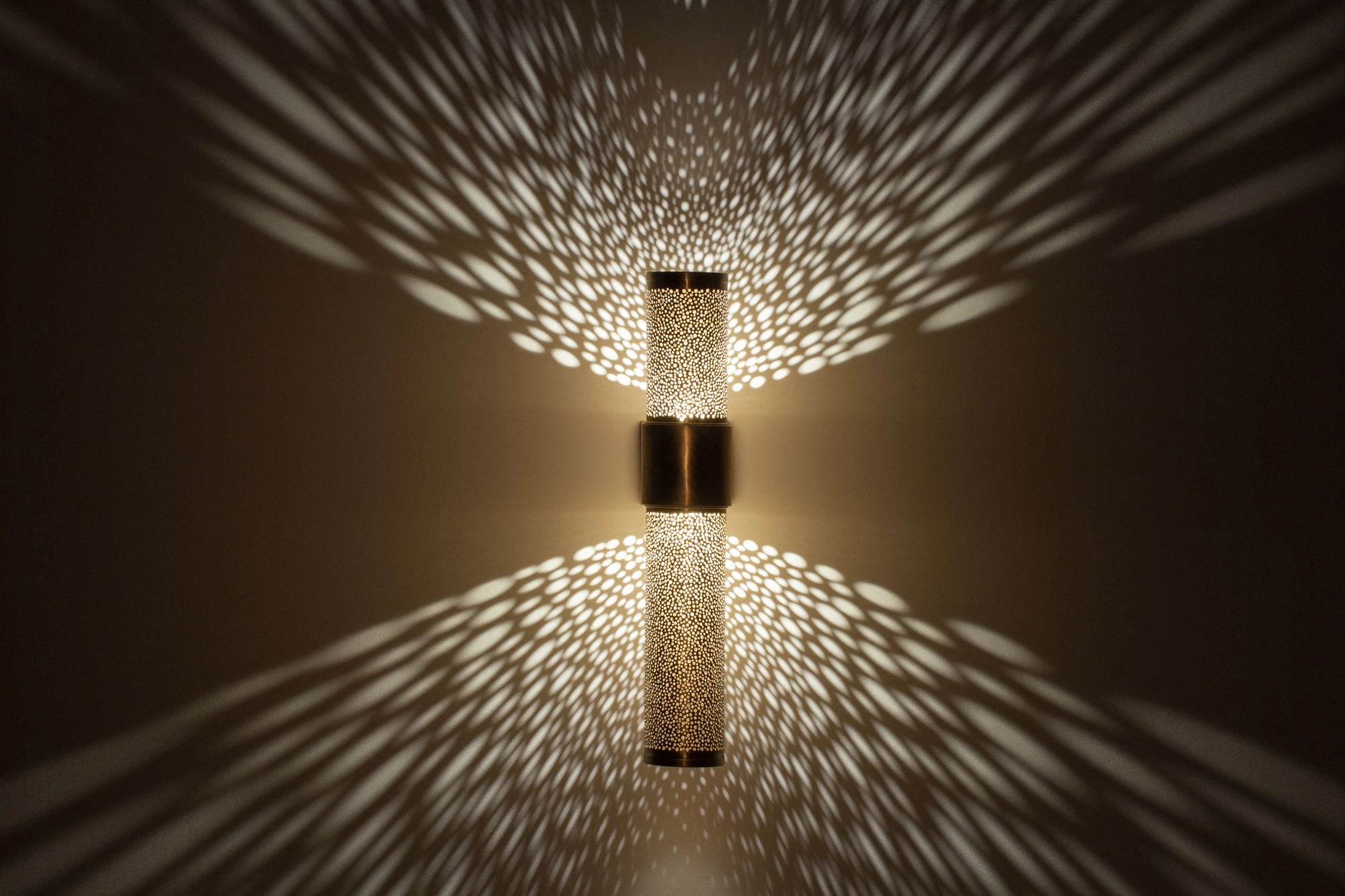 Pierced-brass cropped-cylinder wall sconce glowing, casting a symmetrical burst of light up and down the wall.