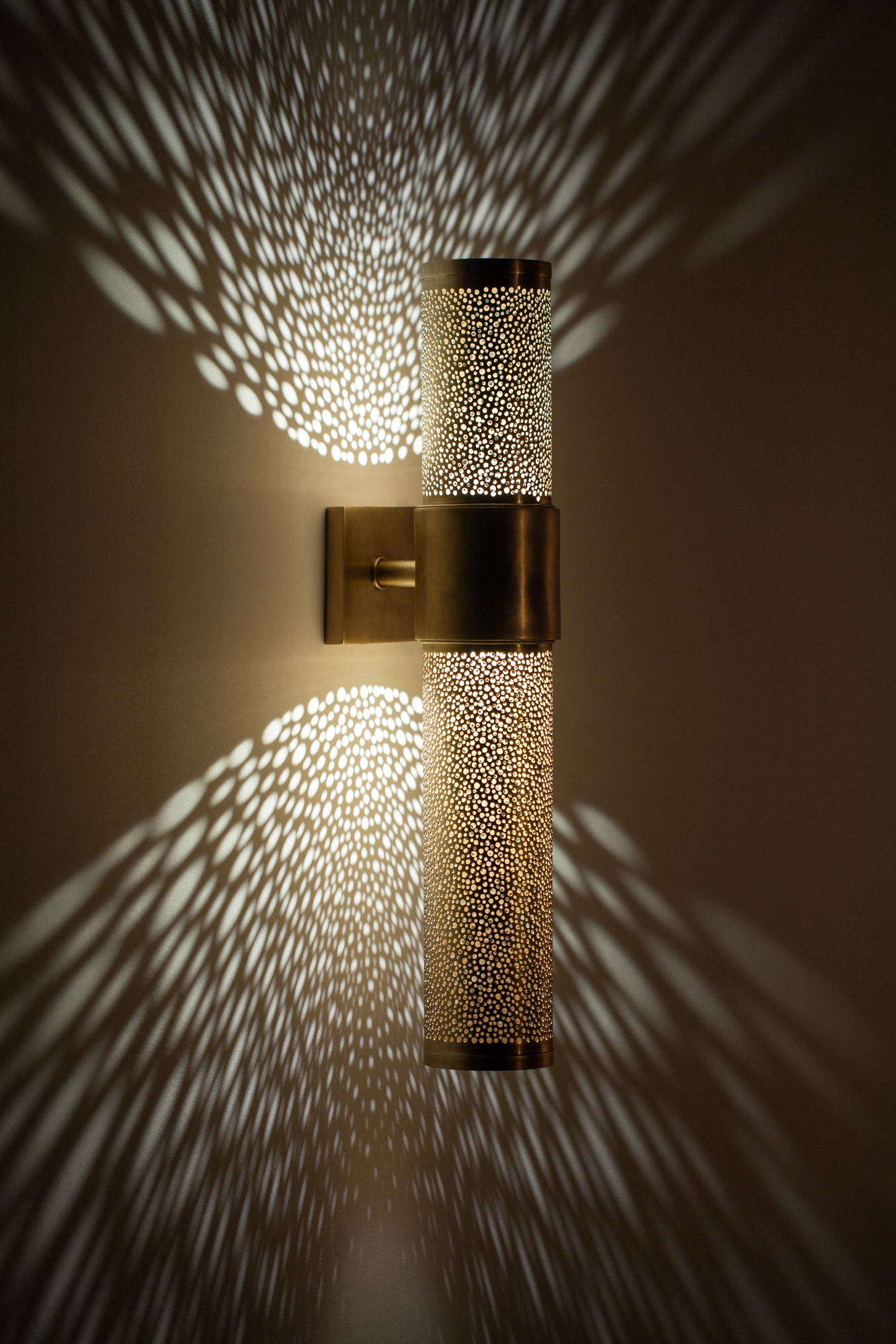 Tall pierced-brass cropped-cylinder wall light glowing, scattering feathery rays of light across the wall.