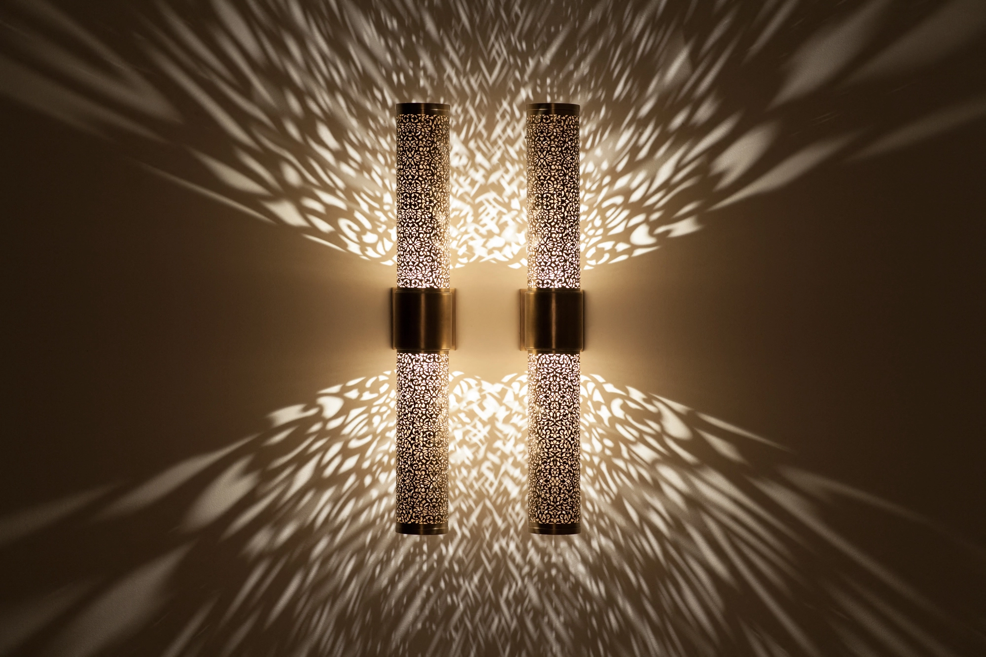 Two pierced-brass cylinder wall lights glowing close together, scattering soft rays of light over the wall.