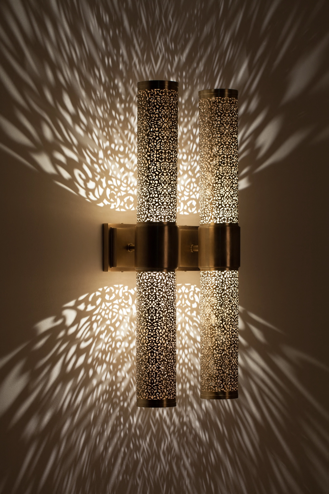 Pair of pierced-brass cylinder wall lights glowing side by side, throwing a dense feathered light burst across the wall.