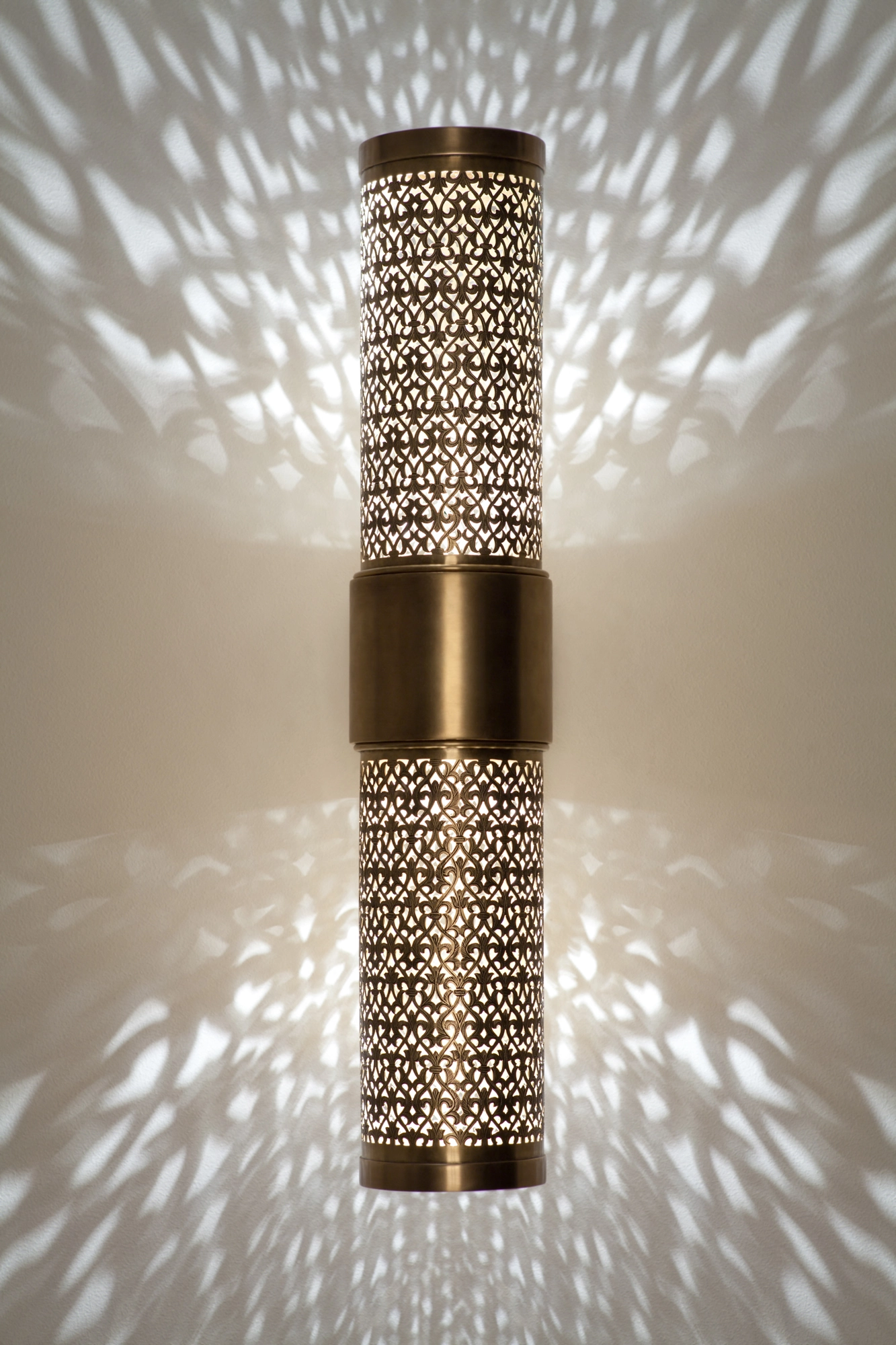 Pierced-brass cylinder wall light glowing on a bracket, casting a bright feathered halo across the wall.
