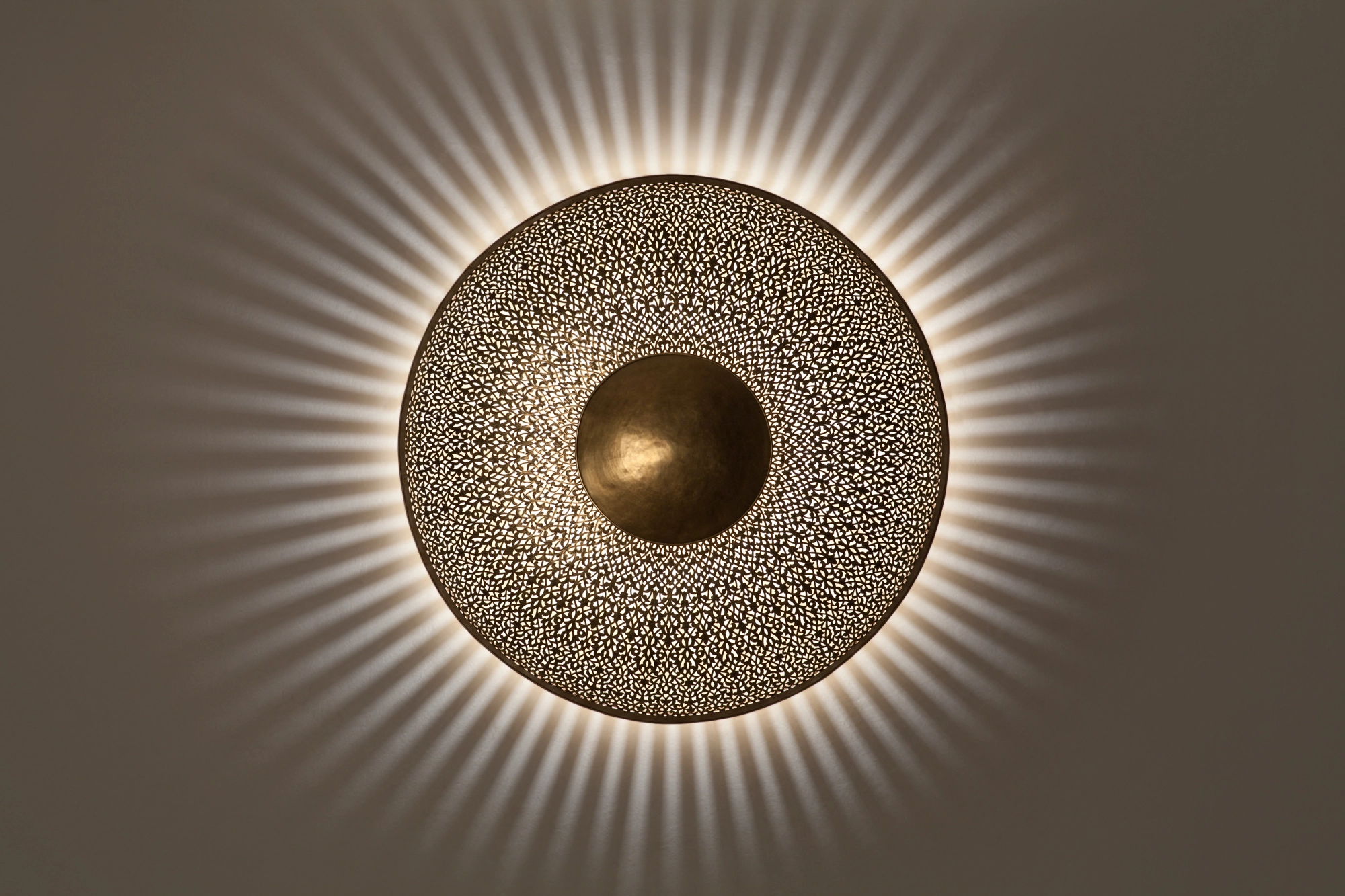 Round pierced-brass sunburst wall light with a domed center, casting a soft radiating halo of light on the wall.