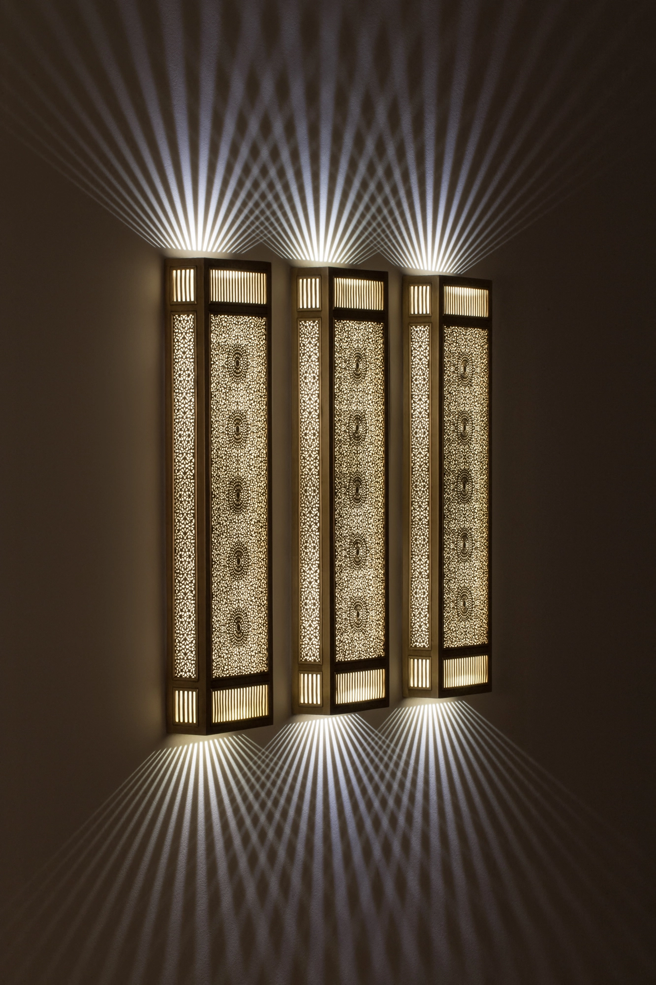 Triple vertical pierced-brass rectangular box wall light glowing, casting upward and downward fans of light.