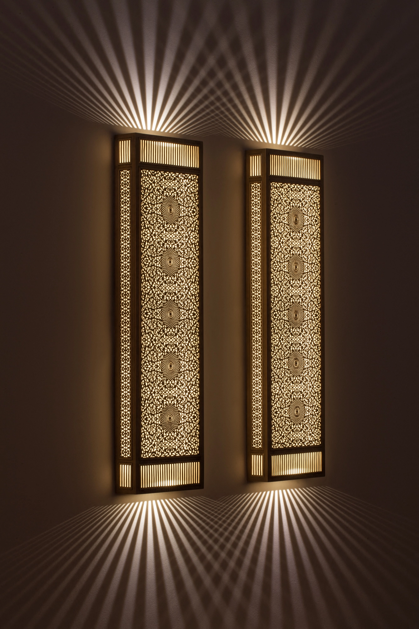 Pair of pierced-brass rectangular box wall lights glowing side by side, throwing star-like rays up and down a dark wall.
