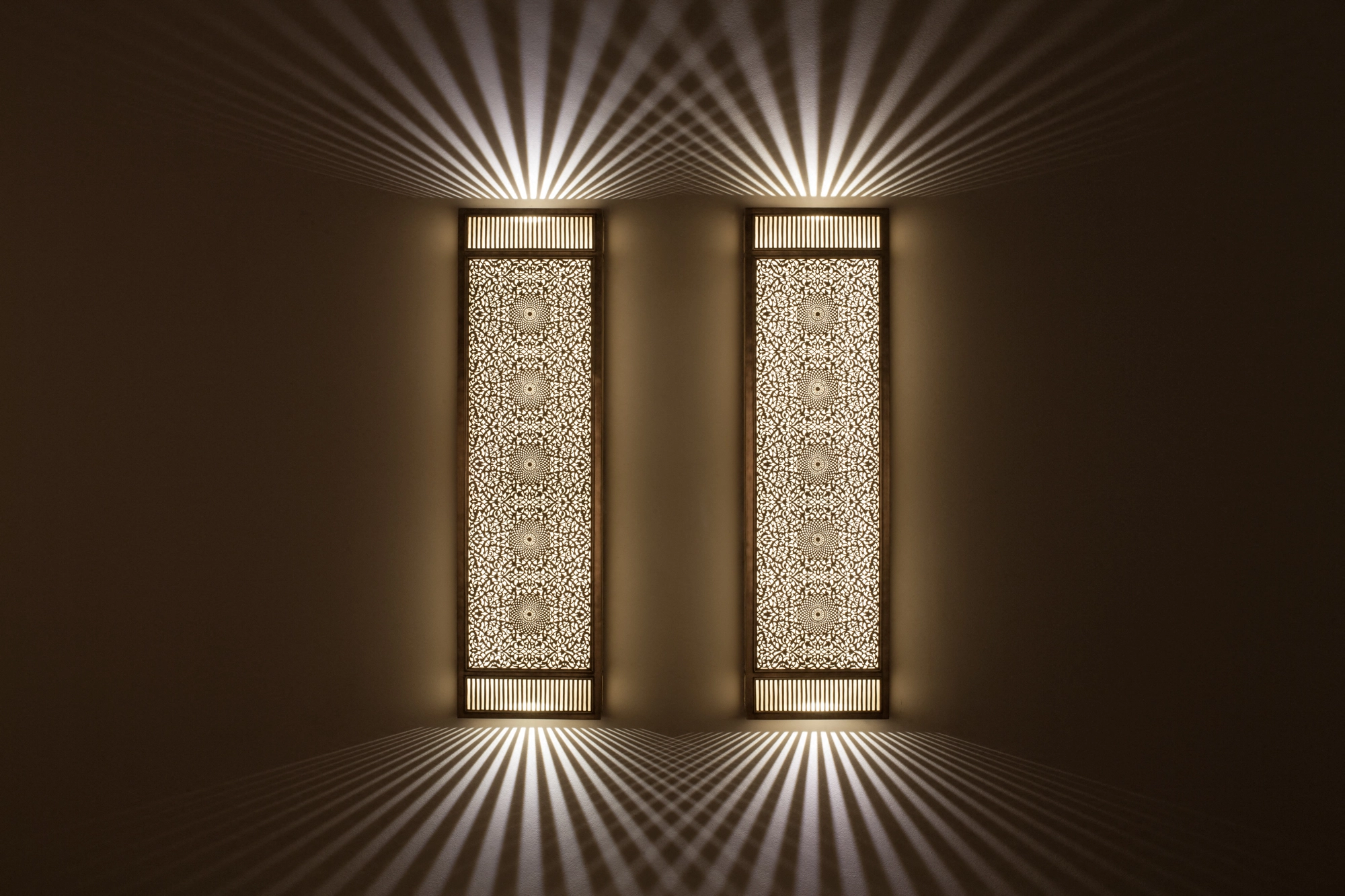 Two slim pierced-brass rectangular box wall lights glowing on a dark wall, casting symmetrical up-and-down light rays.