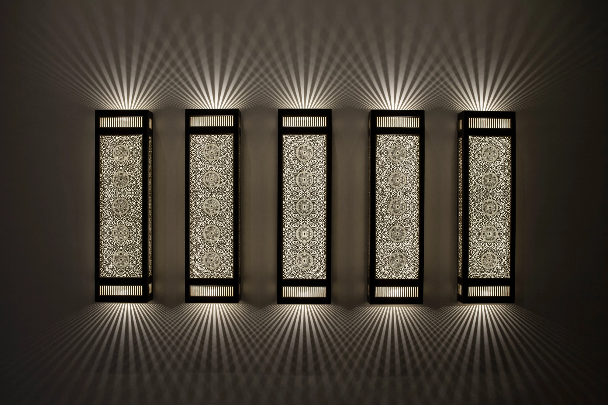 Row of five small pierced-brass rectangular box wall lights glowing softly against a dark wall.