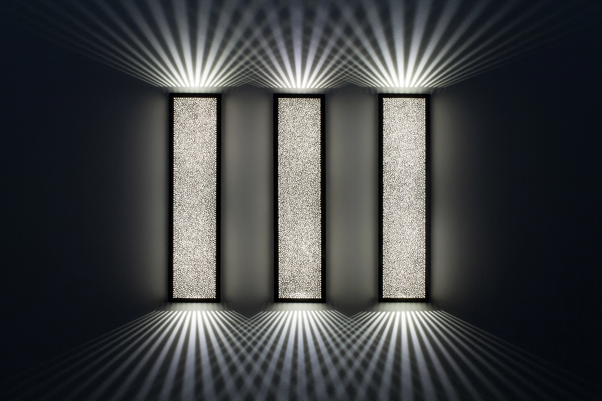 Three pierced-brass rectangular box wall lights glowing in a row, casting fine up-and-down light rays on a dark wall.