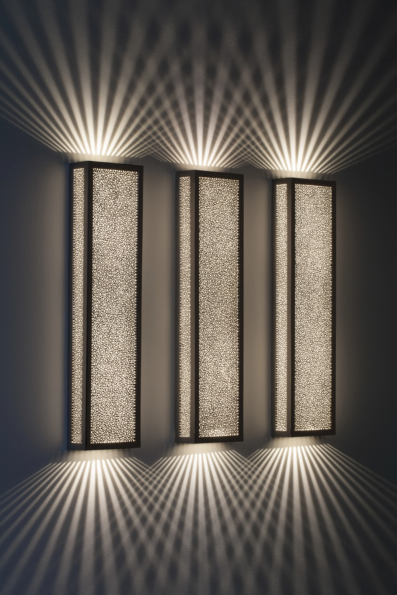 Three offset pierced-brass rectangular box wall lights glowing, throwing a wide starburst of rays up and down.