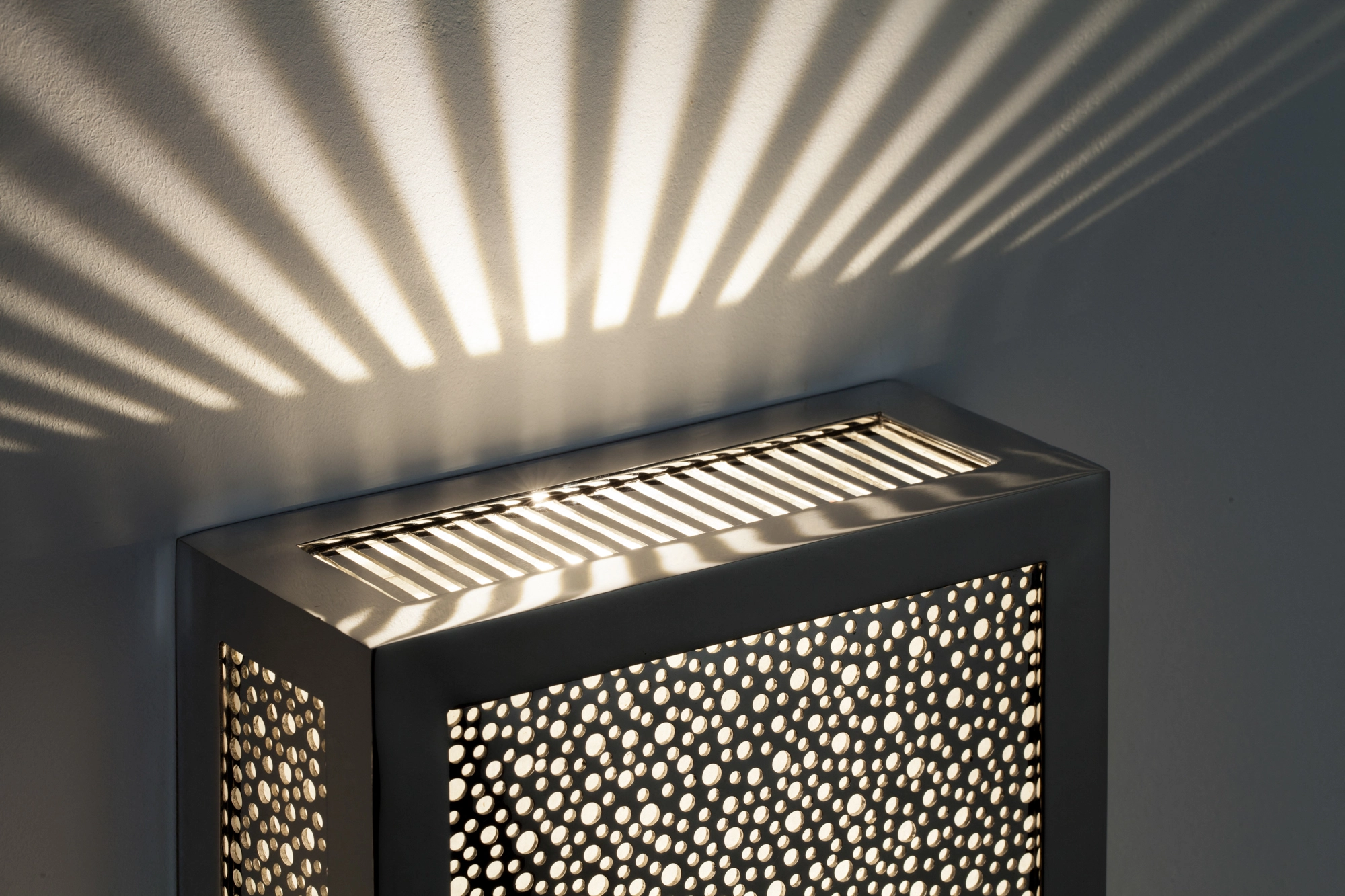 Pierced-brass rectangular box wall light seen at an angle, casting an upward fan of light and dotted shadows.