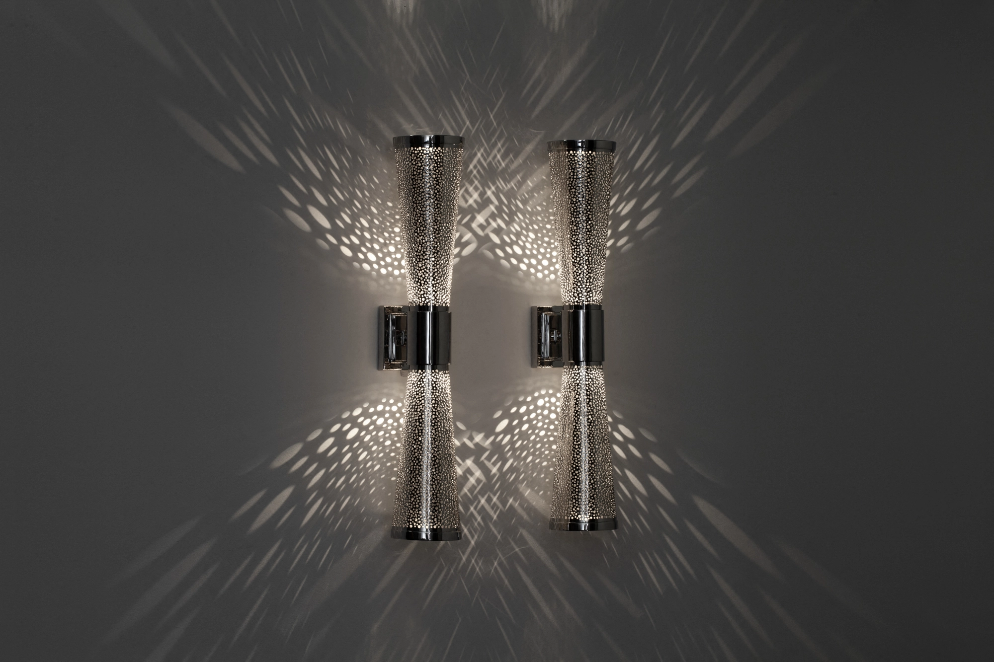 Twin pierced cone wall sconce with points meeting at center, casting faint feathered light up and down a grey wall.